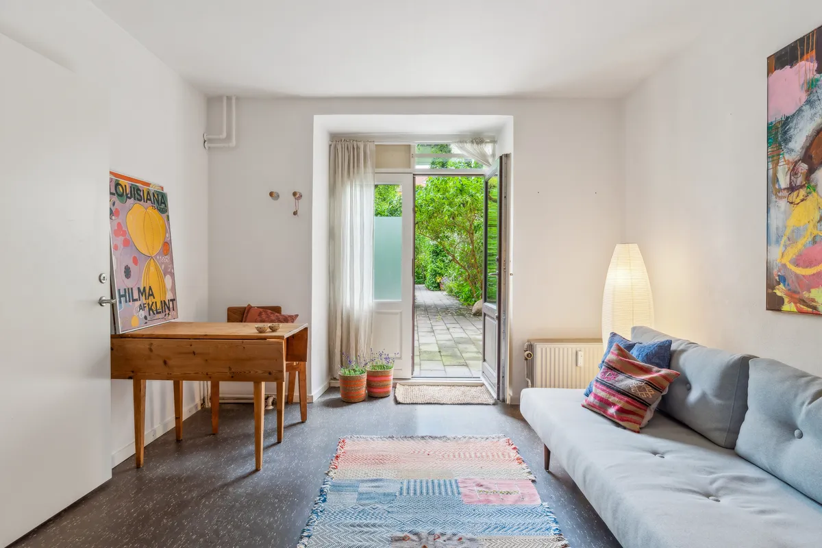 Lejlighed med sofa, skrivebord, plantepotter og maleri. Åben dør med gardin og udgang til baghaven. Lamper og kunstværk på væggen