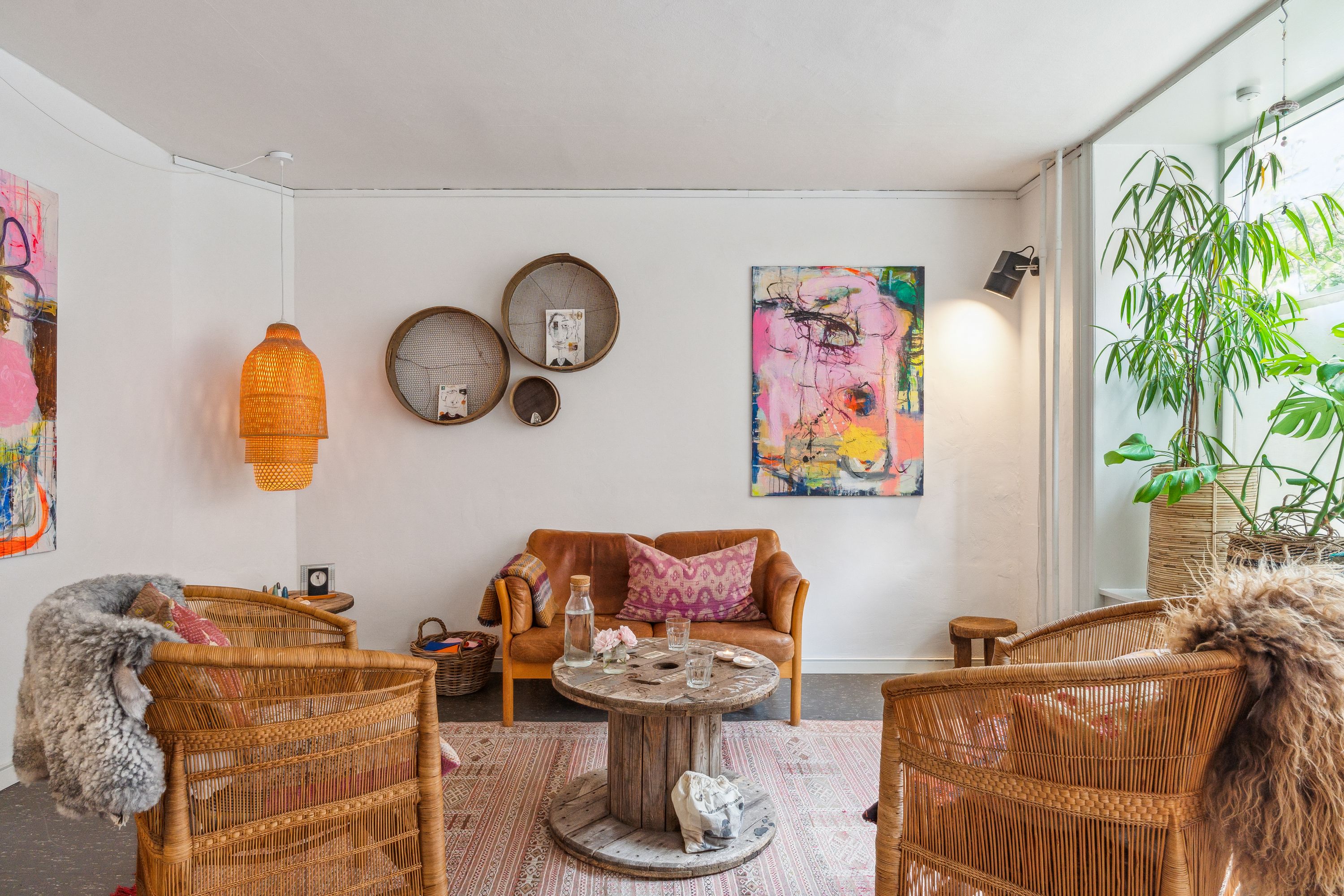 Inde i en indbydende stue med lyse vægge og lysekroner hænger kunstværker og kurve på væggen. Der er en sofa med puder og et rundt træbord i midten af stuen