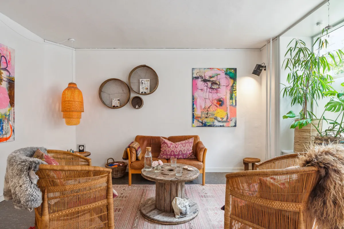 Inde i en indbydende stue med lyse vægge og lysekroner hænger kunstværker og kurve på væggen. Der er en sofa med puder og et rundt træbord i midten af stuen