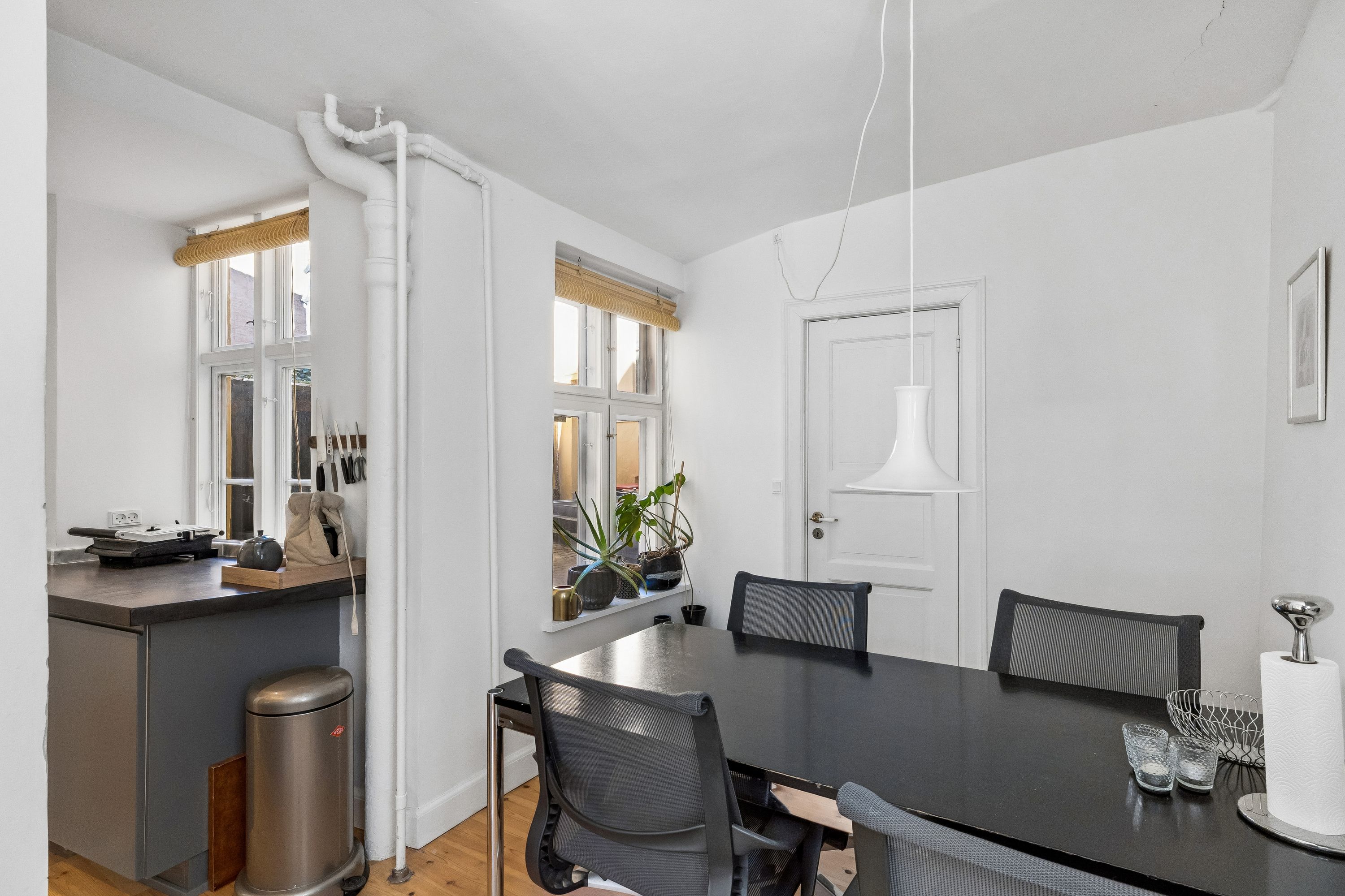 Moderne køkken med køkkenapparater og vindue med plante, samt spisebord med stole og lysekroner i minimalistisk interiør.