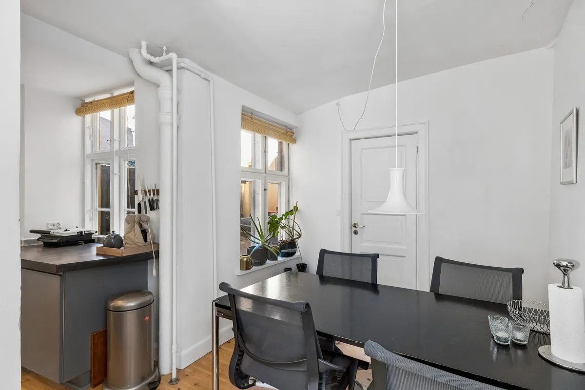 Moderne køkken med køkkenapparater og vindue med plante, samt spisebord med stole og lysekroner i minimalistisk interiør.