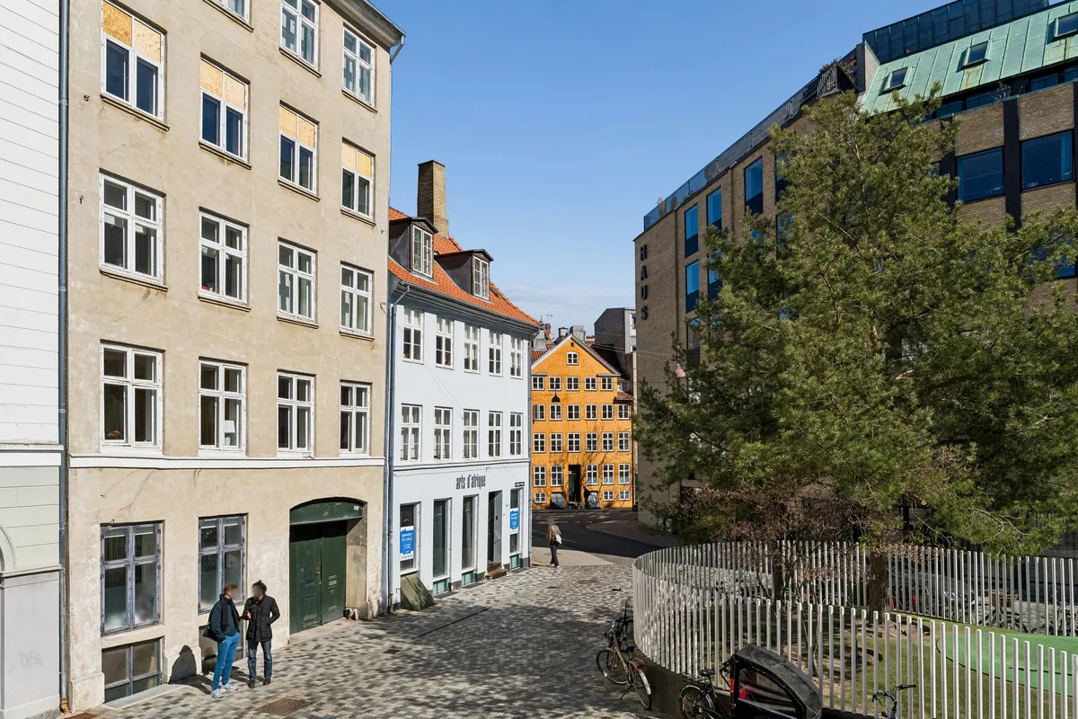 Foto af et kontor i København med træer og huse. To mennesker går på gågaden og går ind i en bygning med røde tag.