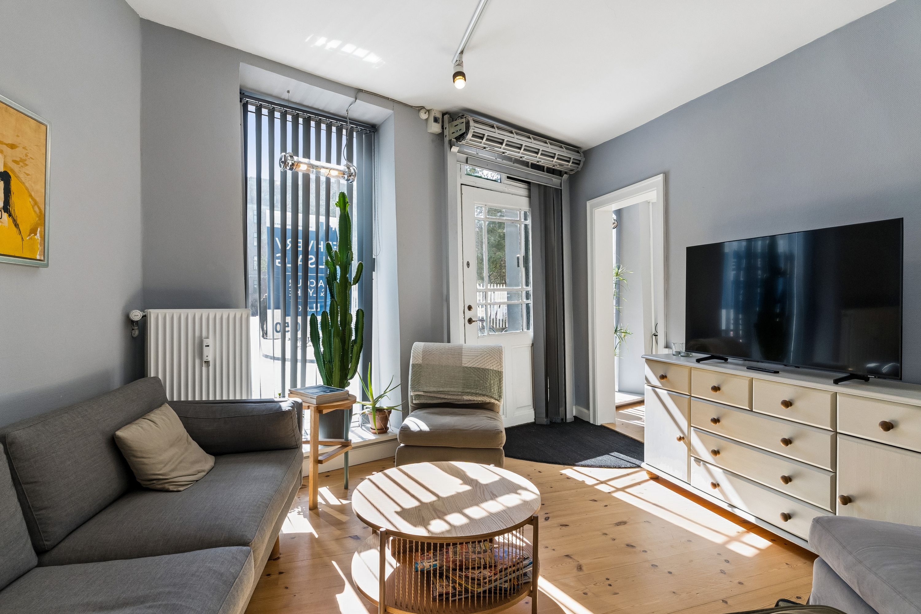 Kontor med sovesofa, tv og hyggeligt møbelsortiment. Solskin trænger ind gennem vinduet og reflekterer på trægulvet.