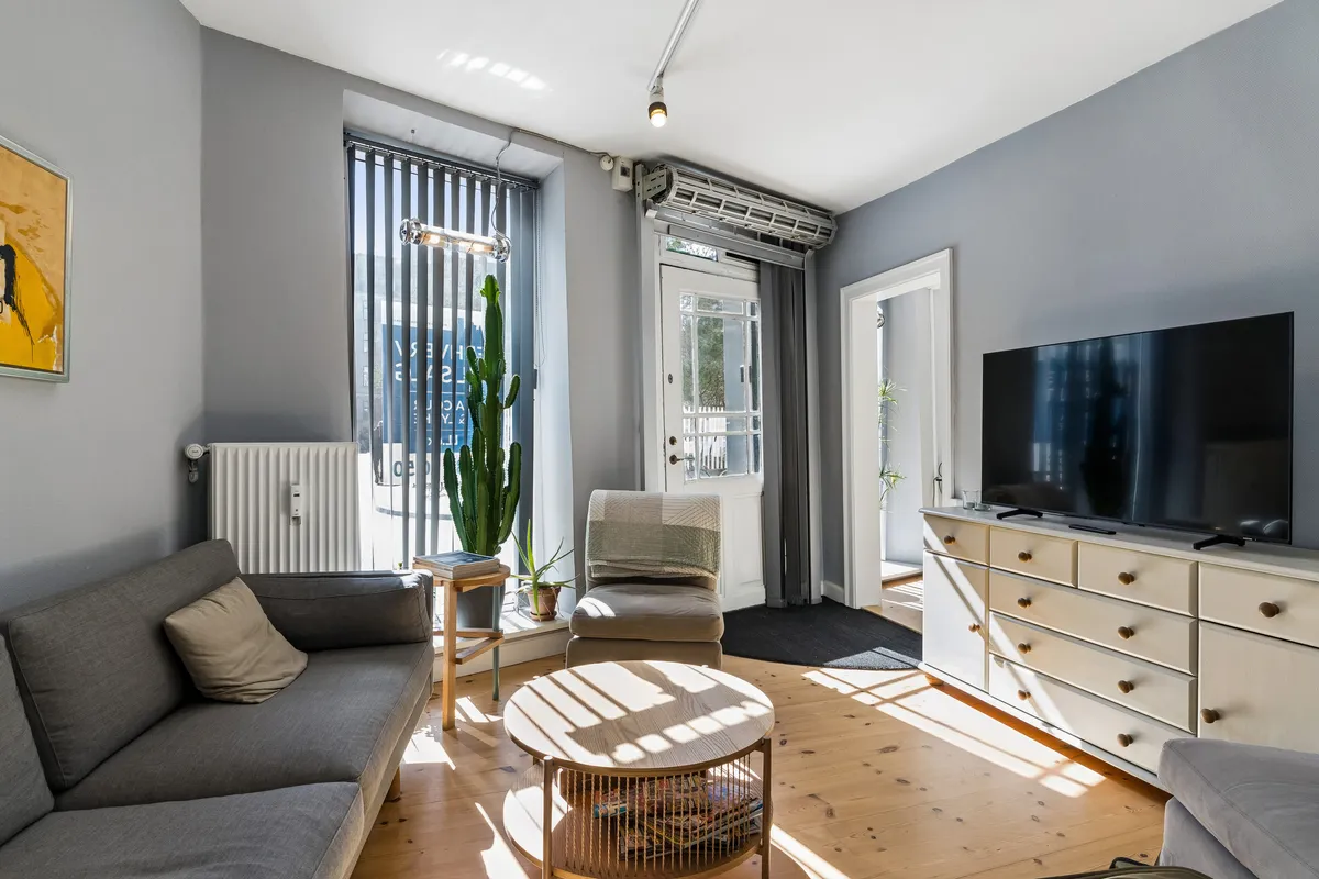 Kontor med sovesofa, tv og hyggeligt møbelsortiment. Solskin trænger ind gennem vinduet og reflekterer på trægulvet.