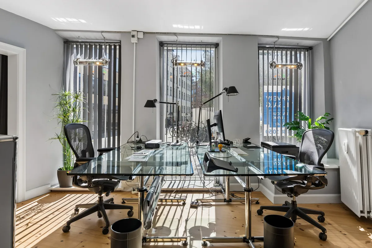 Et moderne kontor med to glasborde, to skrivebordslamper og to kontorstole med hjul samt en skrivebordscomputer og en radiator.