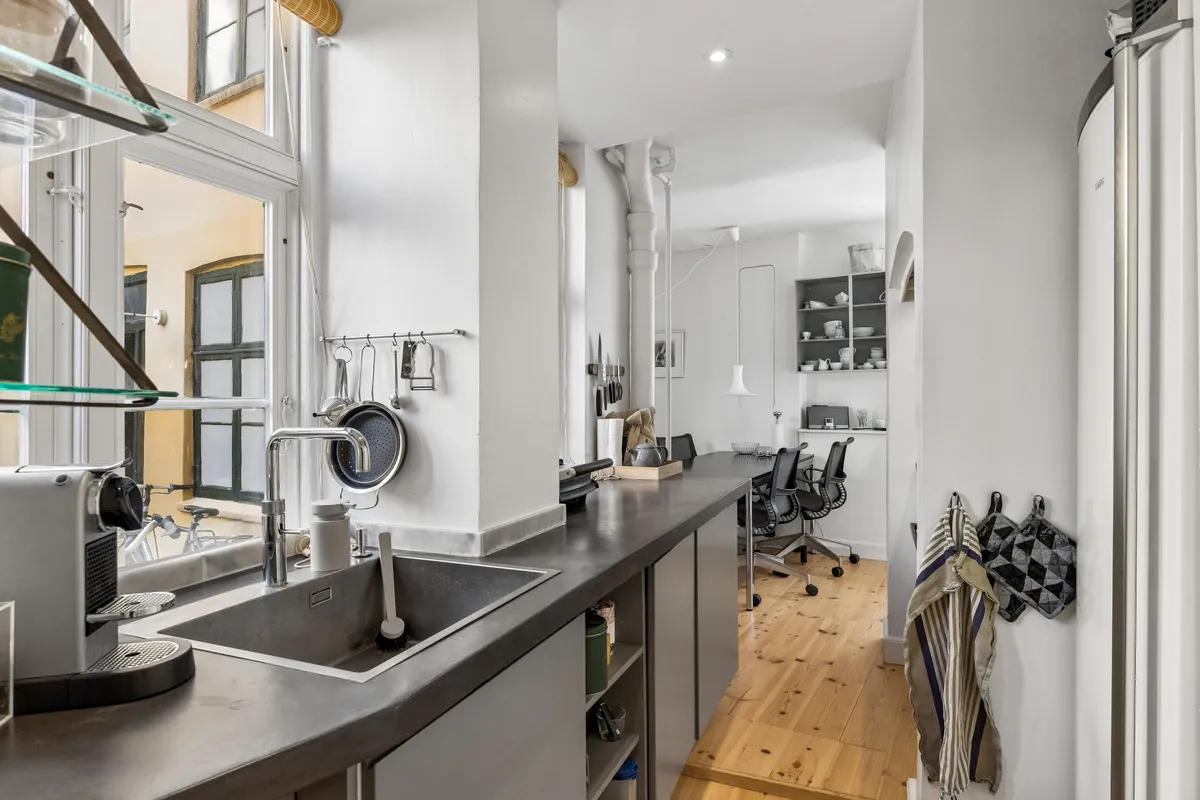 Kontor med lyse lofter og moderne køkken med stålvask og keramikskab. Vinduesglas med refleksioner af byggedelene udenfor.