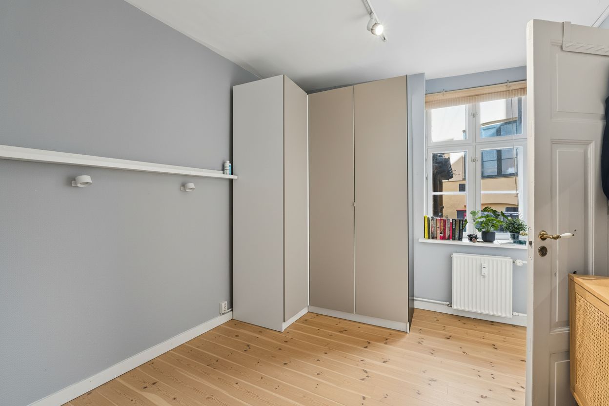 Et tomt kontorlokal med lyse trægulve, to høje garderobeskabe og åbne døre mod vinduet med grønne planter og bøger på vindueskarmen