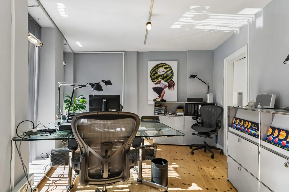 Kontor med skrivebord, stol og plante i stueetagen i København. Væg med tavle og en kvinde. Møbler og teknik placeret i rummet.