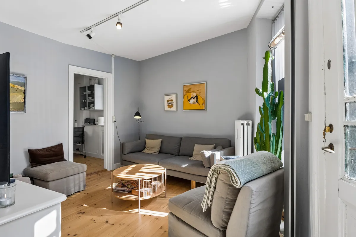 Kontorrum med lyse loftsvinduer og lysegulve. Sofa og hjørneskabsmøbler med puder og dekorationer. Cacti og lamper i rummet.