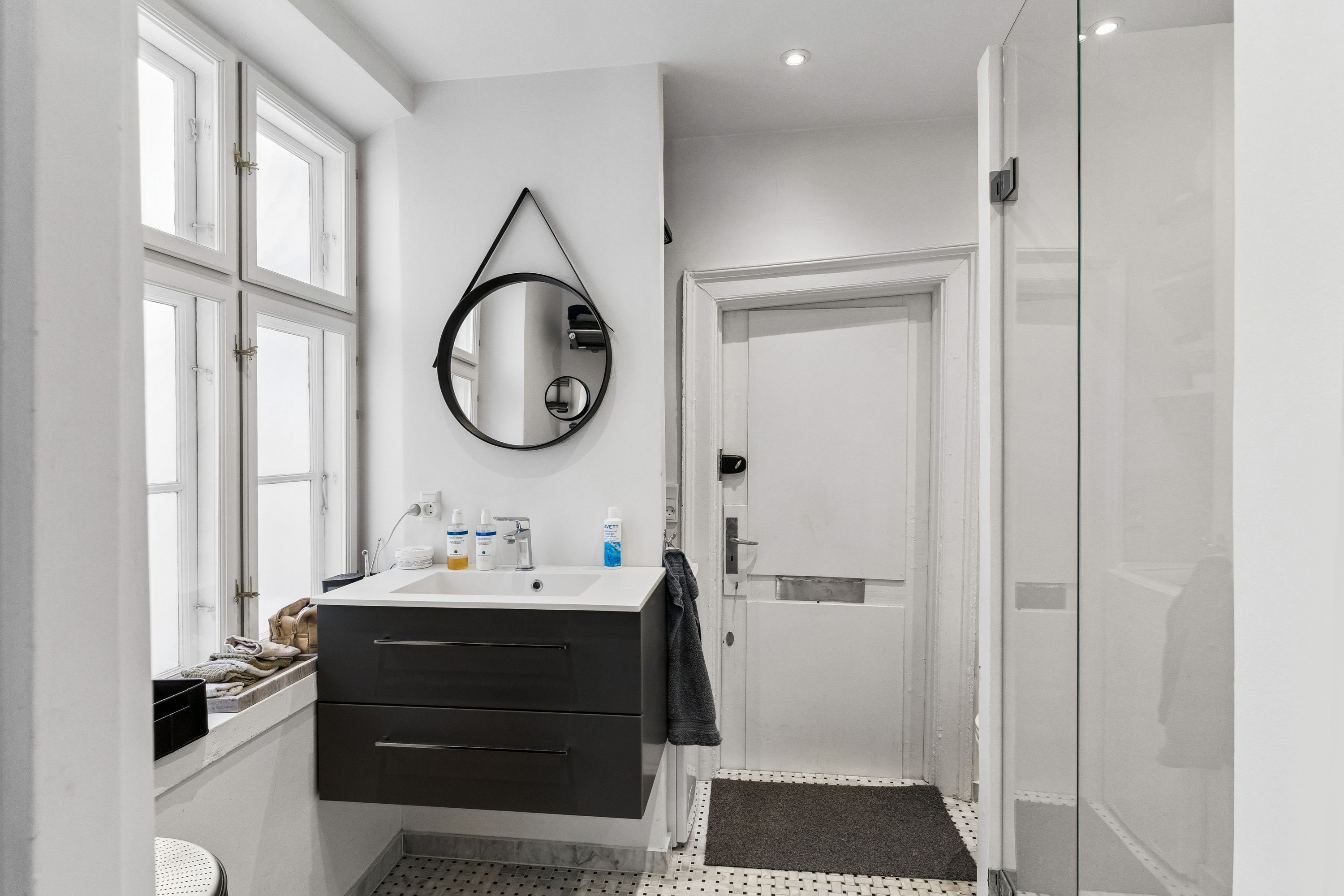 Minimalistisk design med hvidt badeværelse, glasbruser og spejl. Håndklæde og toiletpapir i hvidt. Håndvask med håndklædetørrer og sæbe. Vindue med lysegardiner og udendørsplanter.