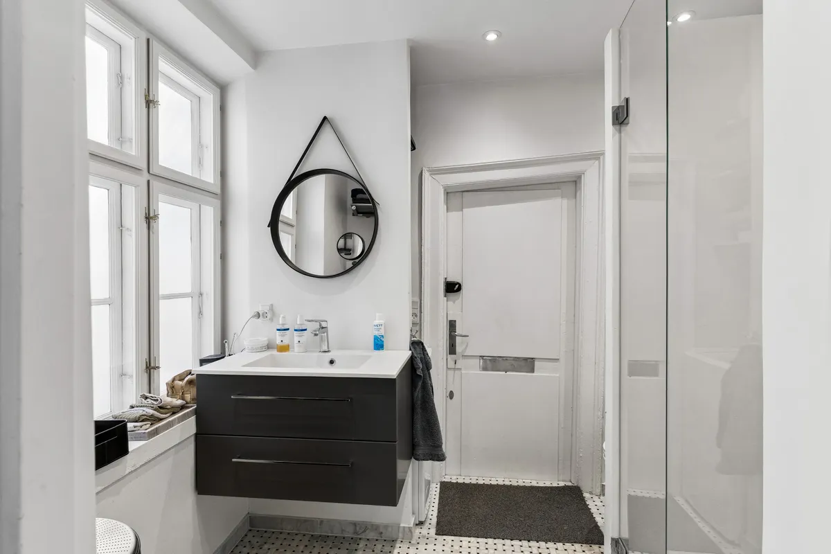 Minimalistisk design med hvidt badeværelse, glasbruser og spejl. Håndklæde og toiletpapir i hvidt. Håndvask med håndklædetørrer og sæbe. Vindue med lysegardiner og udendørsplanter.