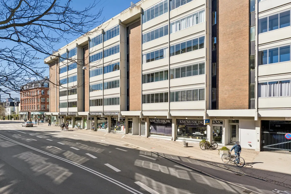 Byggeår 1972. En forretningsbygning med flere butikker på H.C. Ørsteds Vej 1B i Frederiksberg. En mand cykler langs forretningsbygningen.
