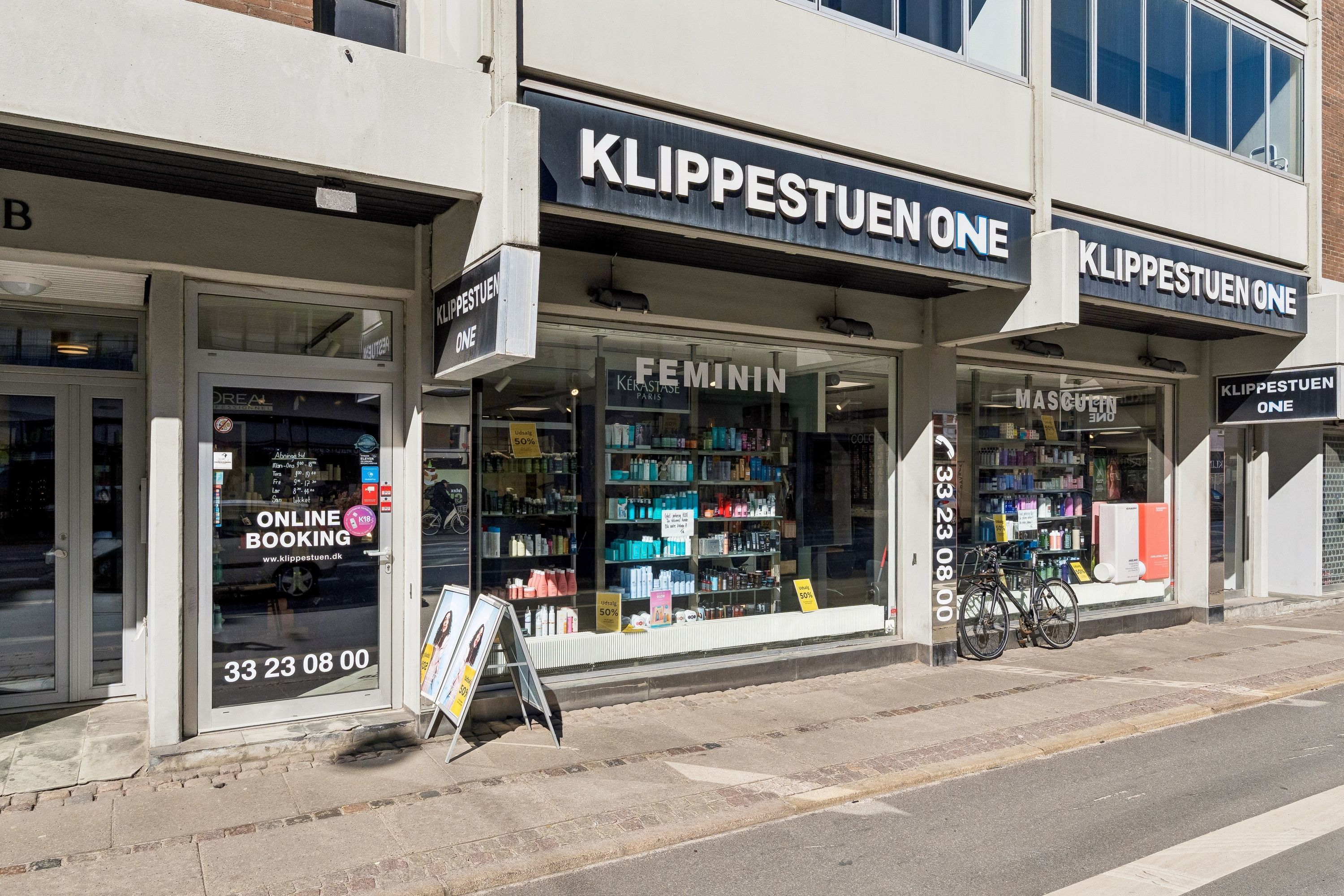 En butik til leje på H.C. Ørsteds Vej 1B, 1879 Frederiksberg. Butikken er indrettet som en frisørforretning med KLIPPESTUEN ONE-skilt og FEMUNIN-produkter i udsigten. Der er også en cykel parkeret udenfor.
