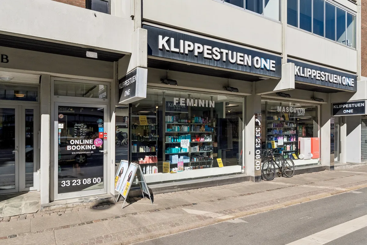 En butik til leje på H.C. Ørsteds Vej 1B, 1879 Frederiksberg. Butikken er indrettet som en frisørforretning med KLIPPESTUEN ONE-skilt og FEMUNIN-produkter i udsigten. Der er også en cykel parkeret udenfor.