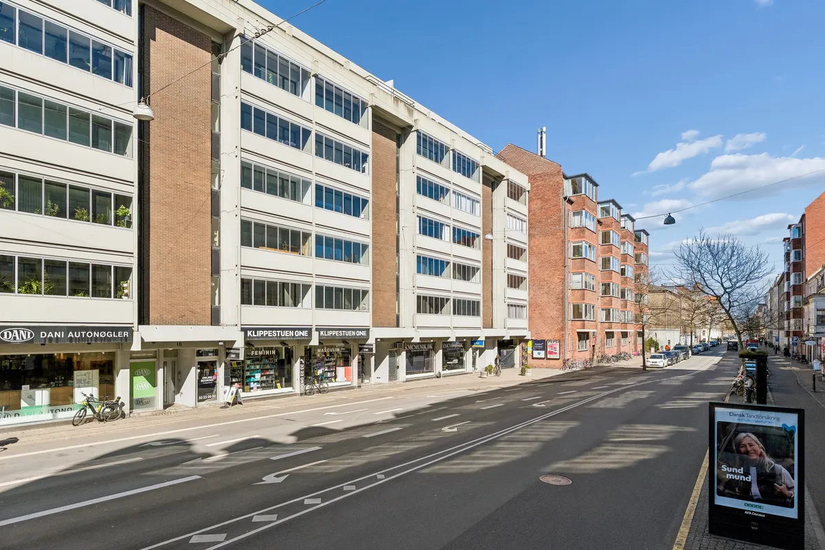 Mennesker går langs en tom gade i Frederiksberg med en frisørforretning og et bibliotek i baggrunden. Vejen er udmærket til cykel- og bilkørsel.