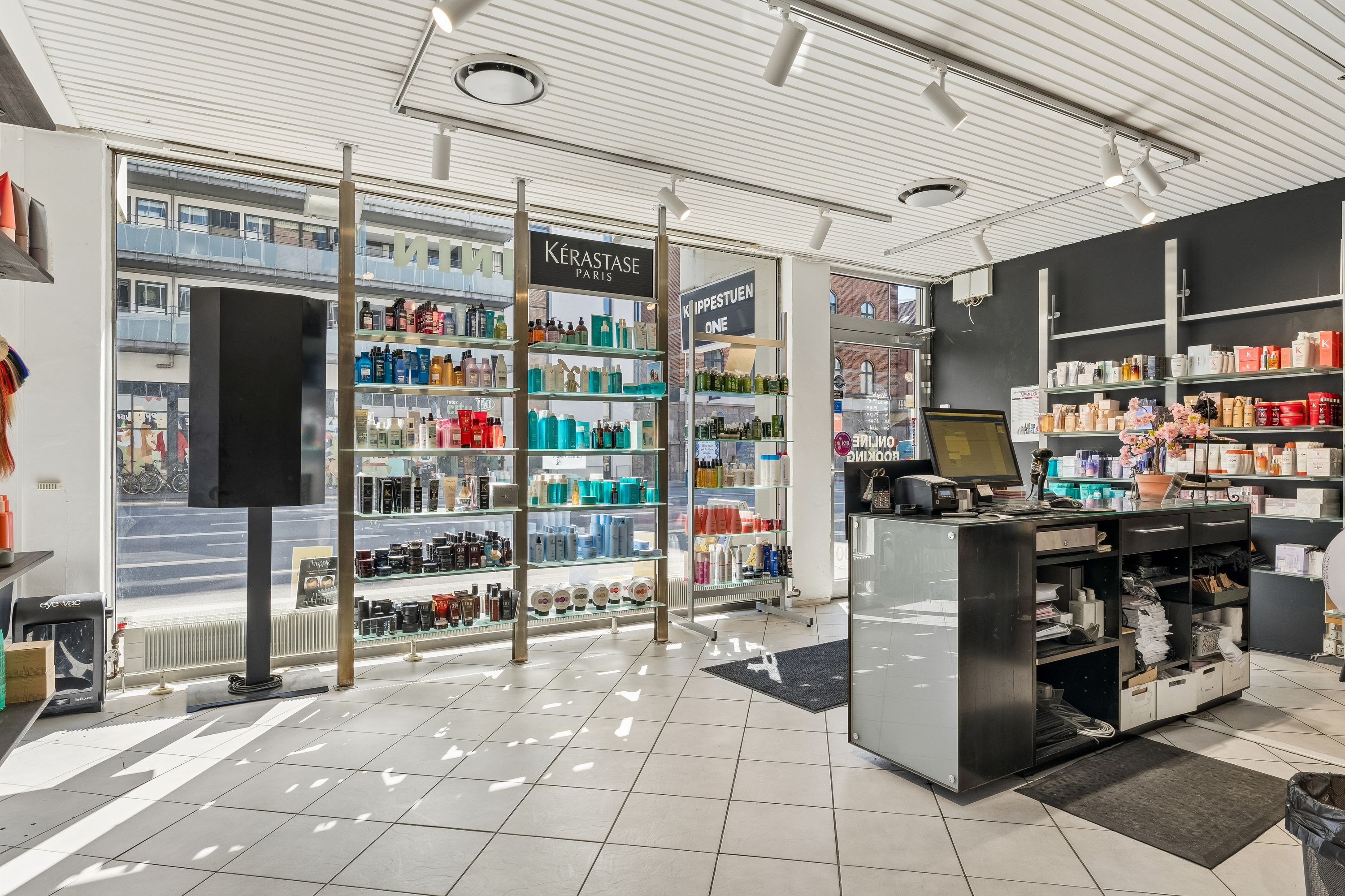 Store frisørforretning med flere skønne produkter i hvidt og blåt udsæt i flere hylder. Butikken har en længe med flere hylder og et skrivebord med en computer og en kasseapparat.