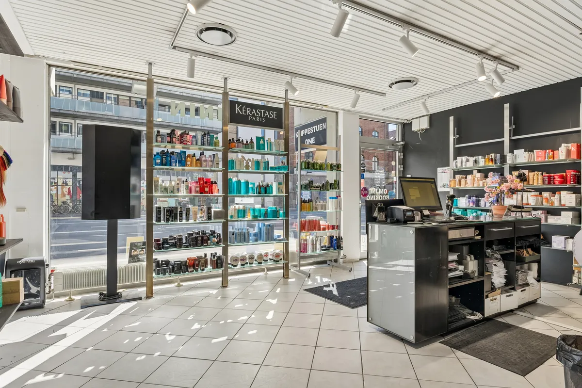 Store frisørforretning med flere skønne produkter i hvidt og blåt udsæt i flere hylder. Butikken har en længe med flere hylder og et skrivebord med en computer og en kasseapparat.