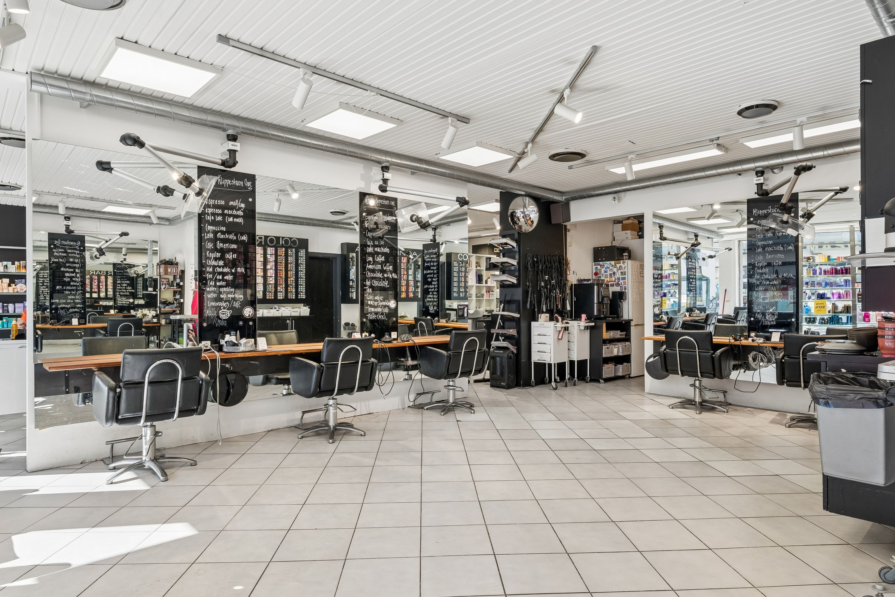 Modernt indrettet frisørsalon med lysegulve og højtloft. Faciliteterne inkluderer hårsæder, hårplejeprodukter og flere smykkeskuffer. Store spejle og hængende lyskatler er installeret