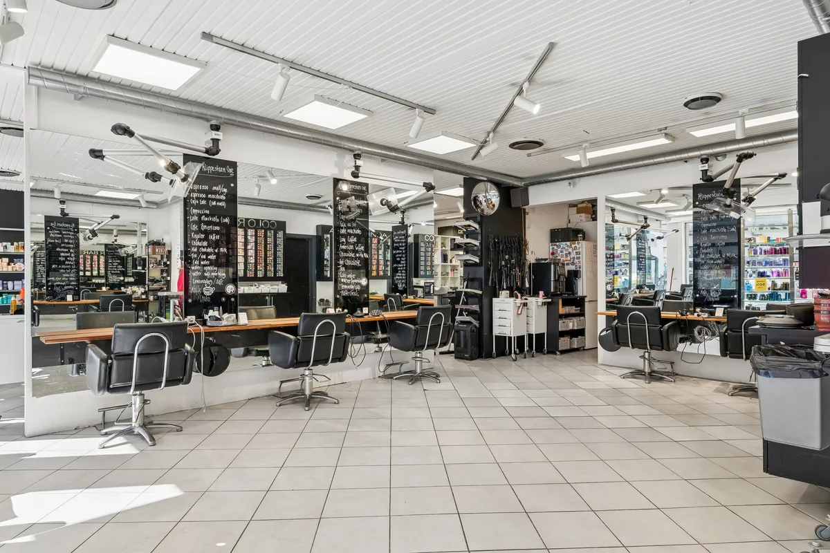 Modernt indrettet frisørsalon med lysegulve og højtloft. Faciliteterne inkluderer hårsæder, hårplejeprodukter og flere smykkeskuffer. Store spejle og hængende lyskatler er installeret