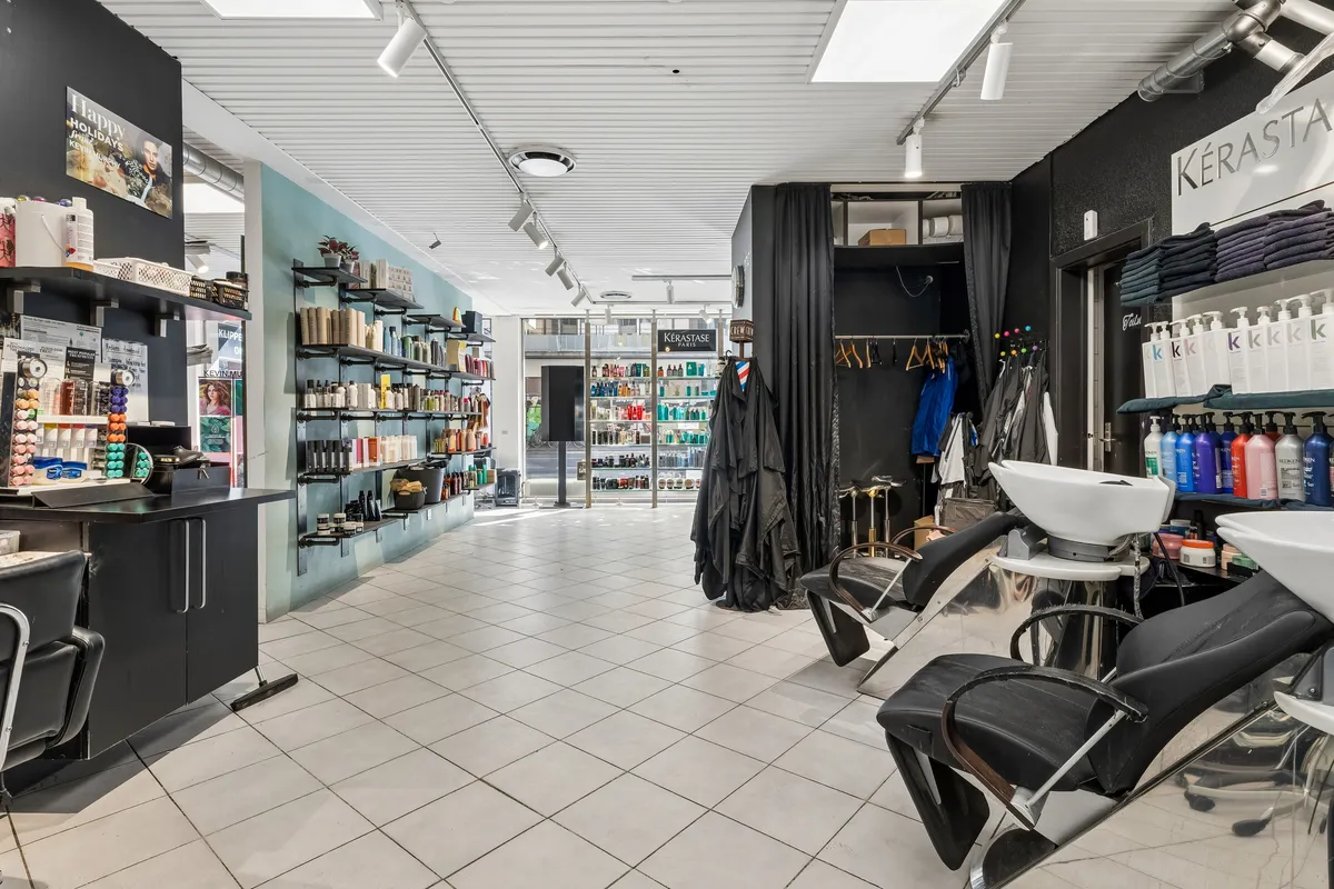 En frisørsalon med lyse loftslampe, moderne udstyr og store lagerhylder med diverse kosmetiske produkter. De to shampoobøsser og stole er placeret i midten af gulvet.