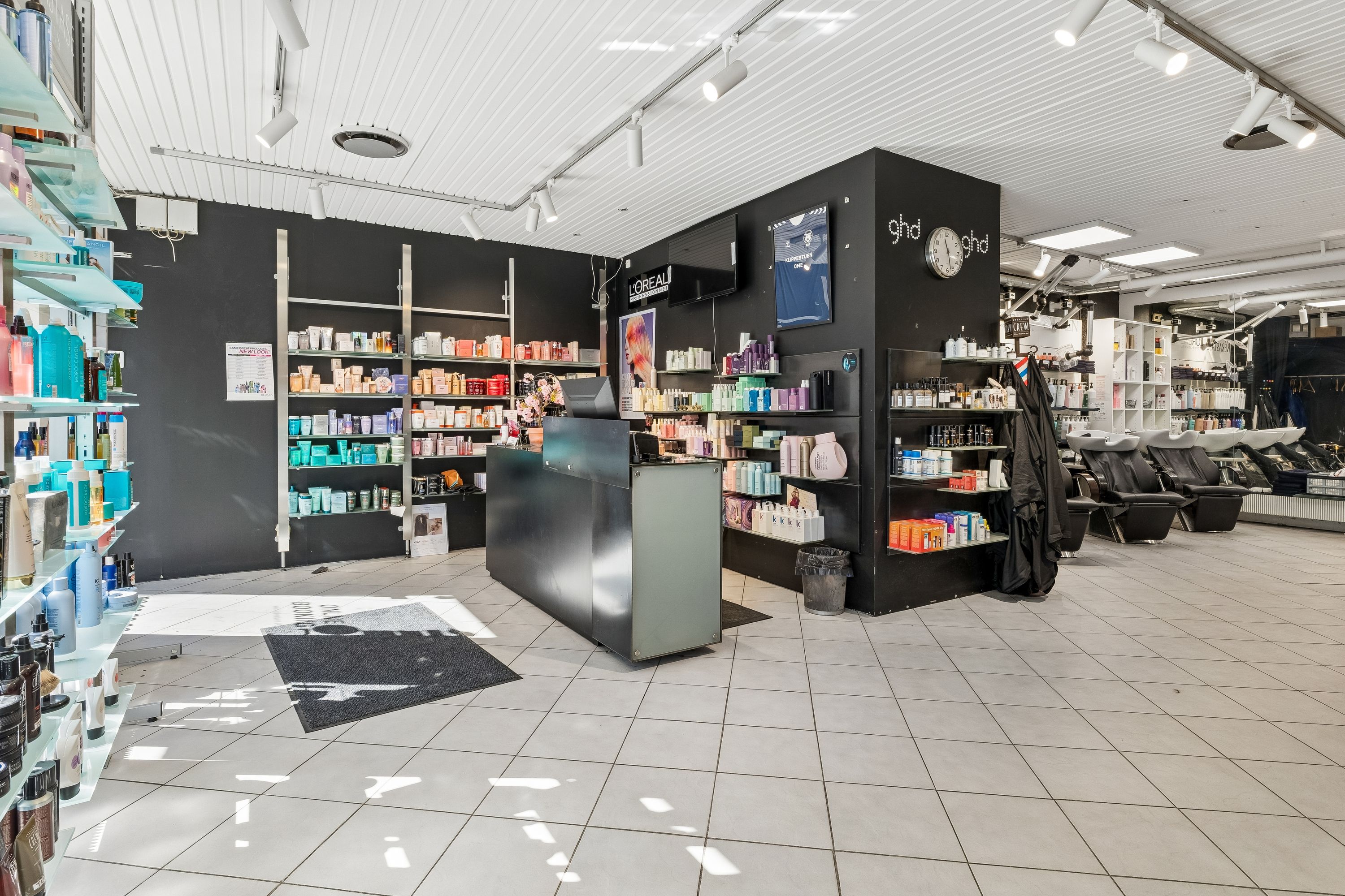 Modern indretning med hvide gulve, lysekroner, høje hylder med kosmetik, og skrivebord med computer. Bagved er en mørk væg med brandnavn og klokke.