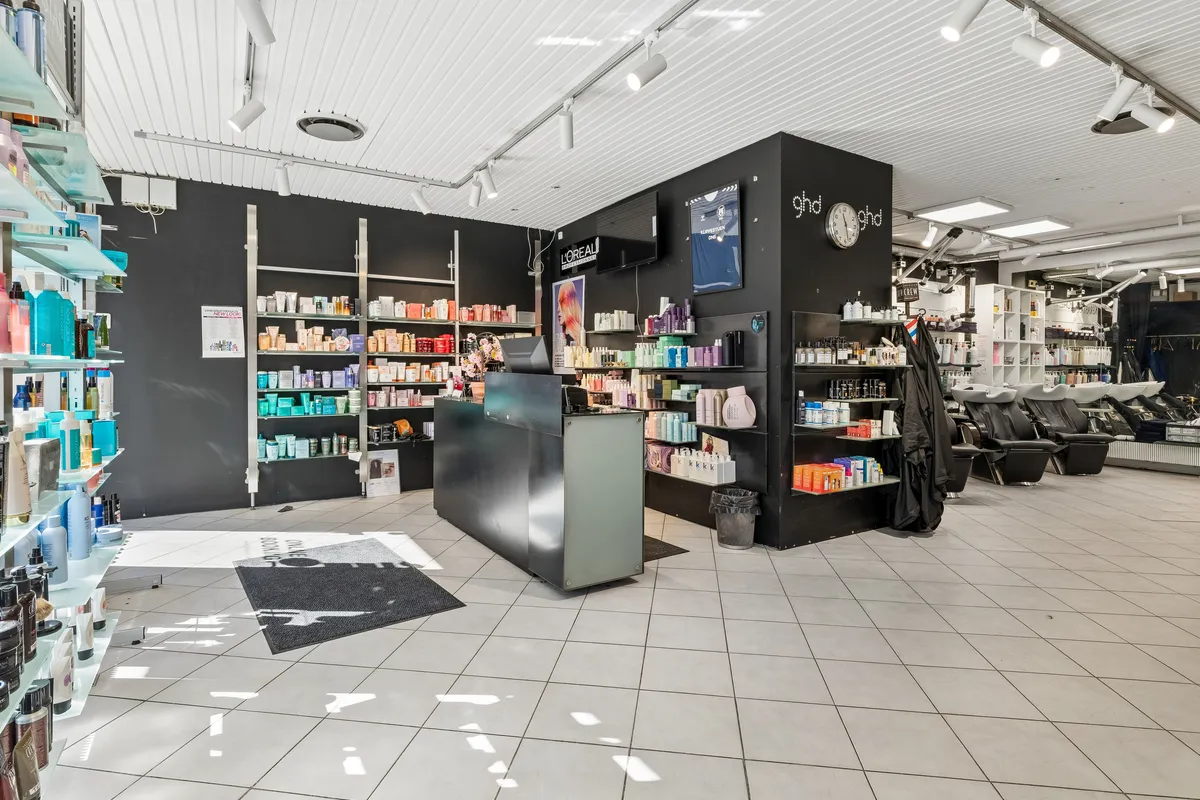 Modern indretning med hvide gulve, lysekroner, høje hylder med kosmetik, og skrivebord med computer. Bagved er en mørk væg med brandnavn og klokke.