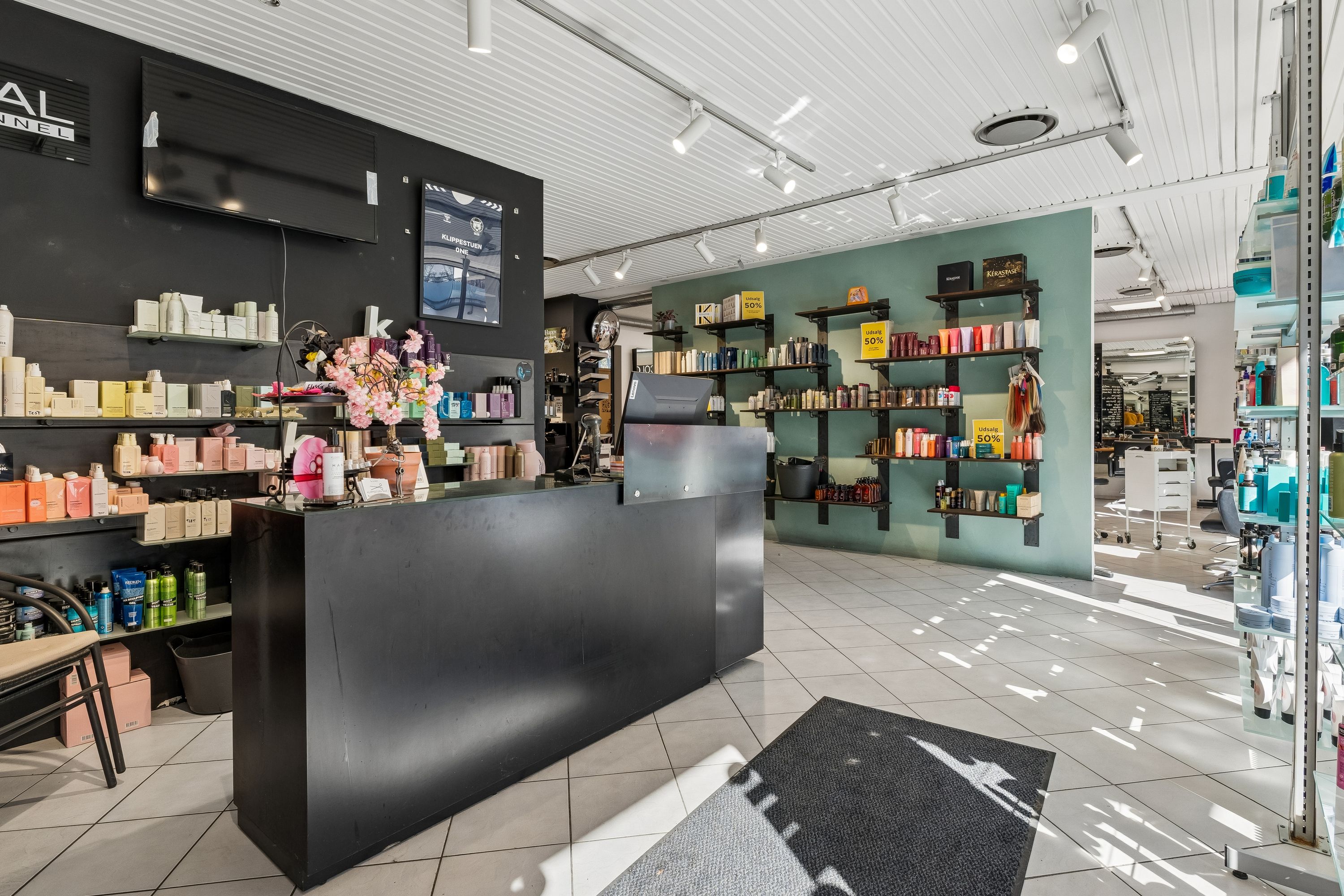 En stor, ren frisørsalon med hvide gulvfliser, moderne møbler og et stort udvalg af hårprodukter og kosmetik på hylderne