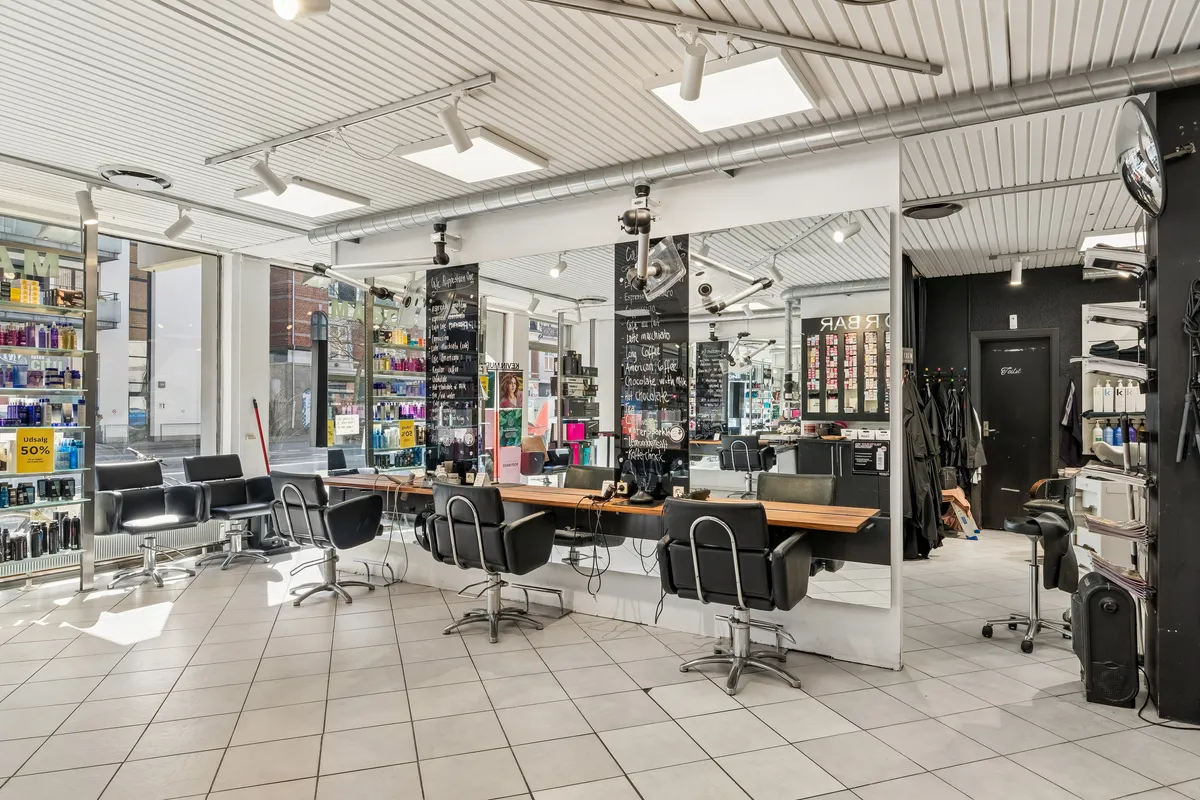 Butik med moderne interiør, højt til loftet, hvidt og sort og lyse hjørner med store spejl. Køkkenbord og flere frisørstole. Produkter i glasskab og et stort sort skab med flasker.