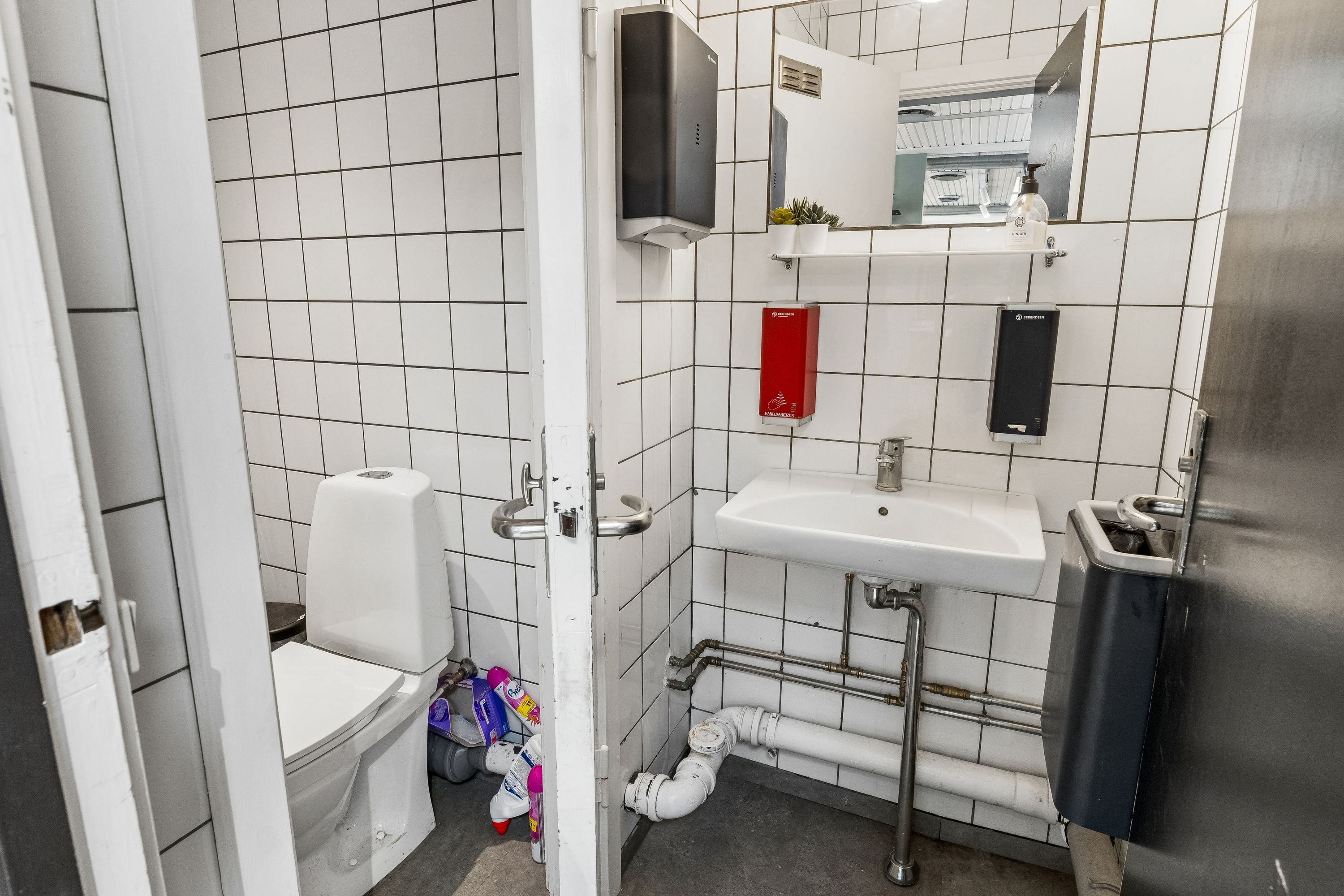 Hvidt toilet i lille toiletrum med lysegrå, firkantede fliser og to håndvask samt hånddesinfektionsstationer på væggen
