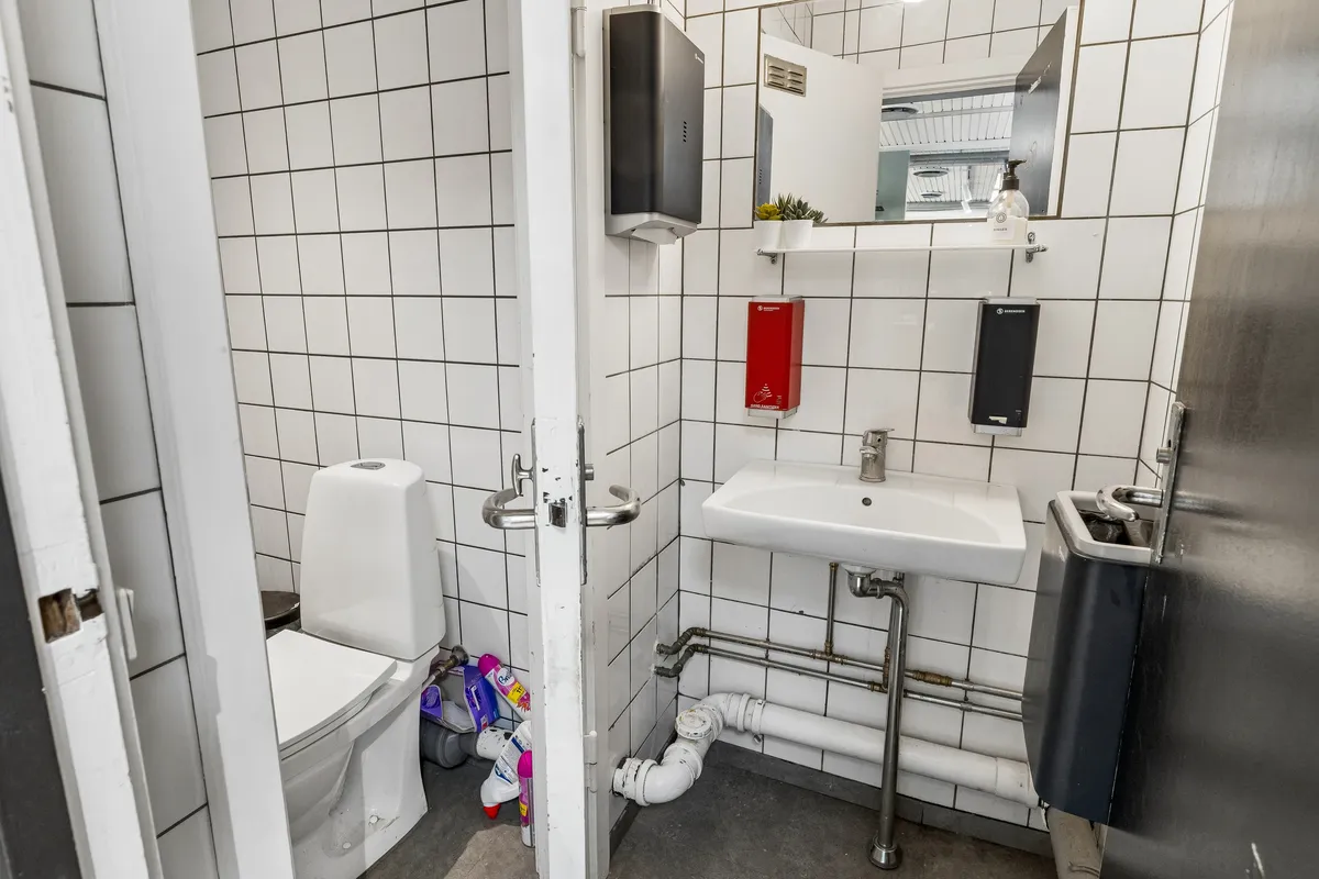 Hvidt toilet i lille toiletrum med lysegrå, firkantede fliser og to håndvask samt hånddesinfektionsstationer på væggen