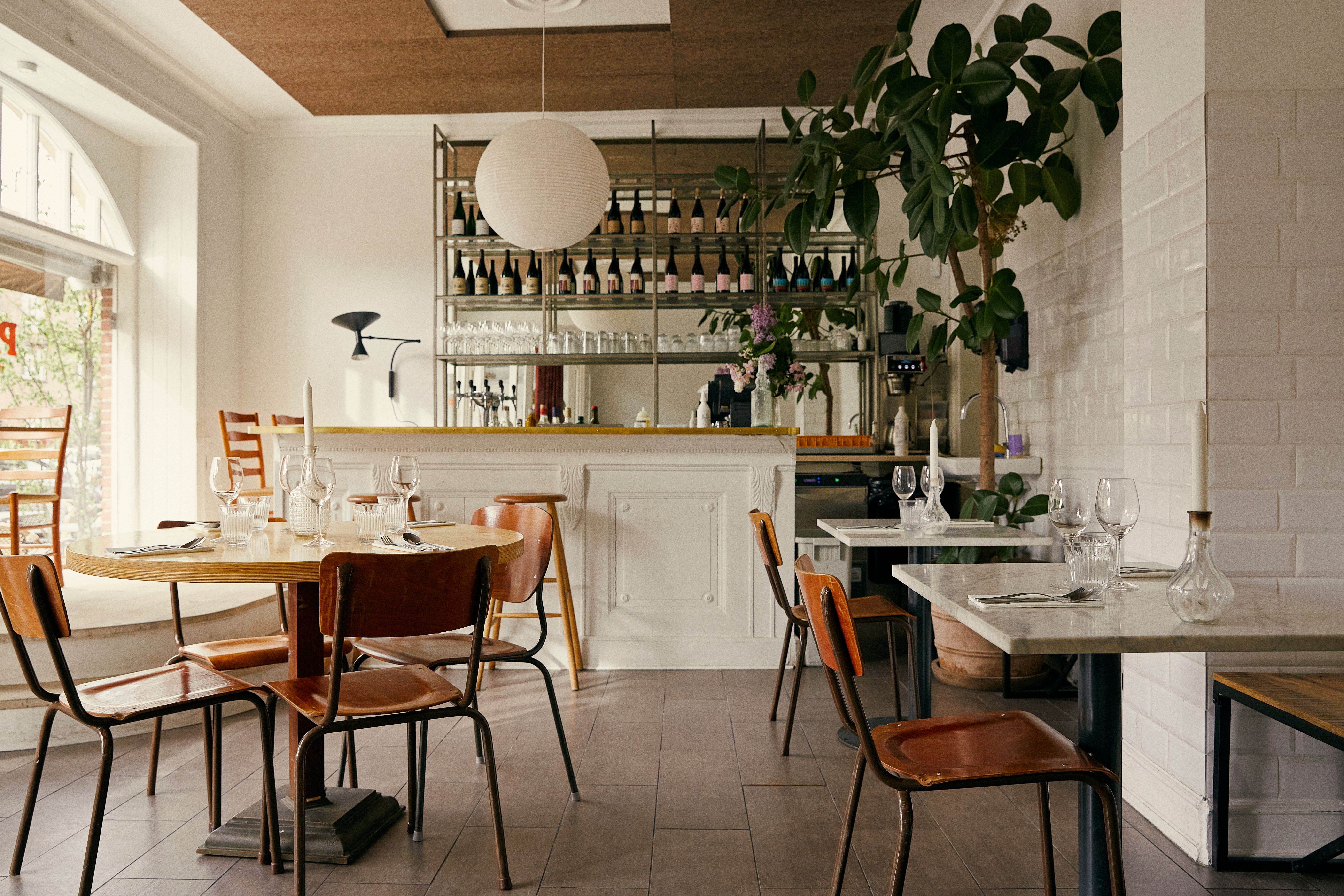 Et moderne og luksuriøst restaurantområde med runde og firkantede borde, lysekroner, smukt grønne planter og glasvinduer.