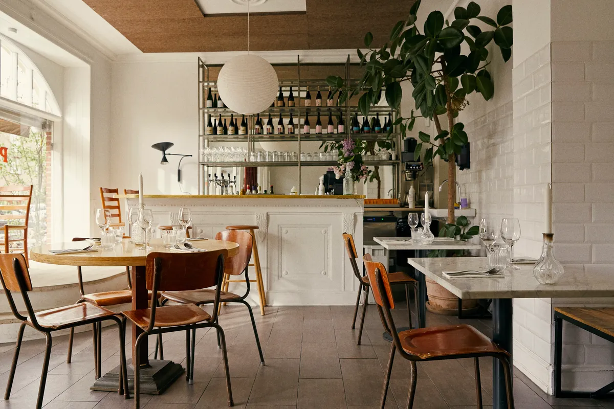 Et moderne og luksuriøst restaurantområde med runde og firkantede borde, lysekroner, smukt grønne planter og glasvinduer.