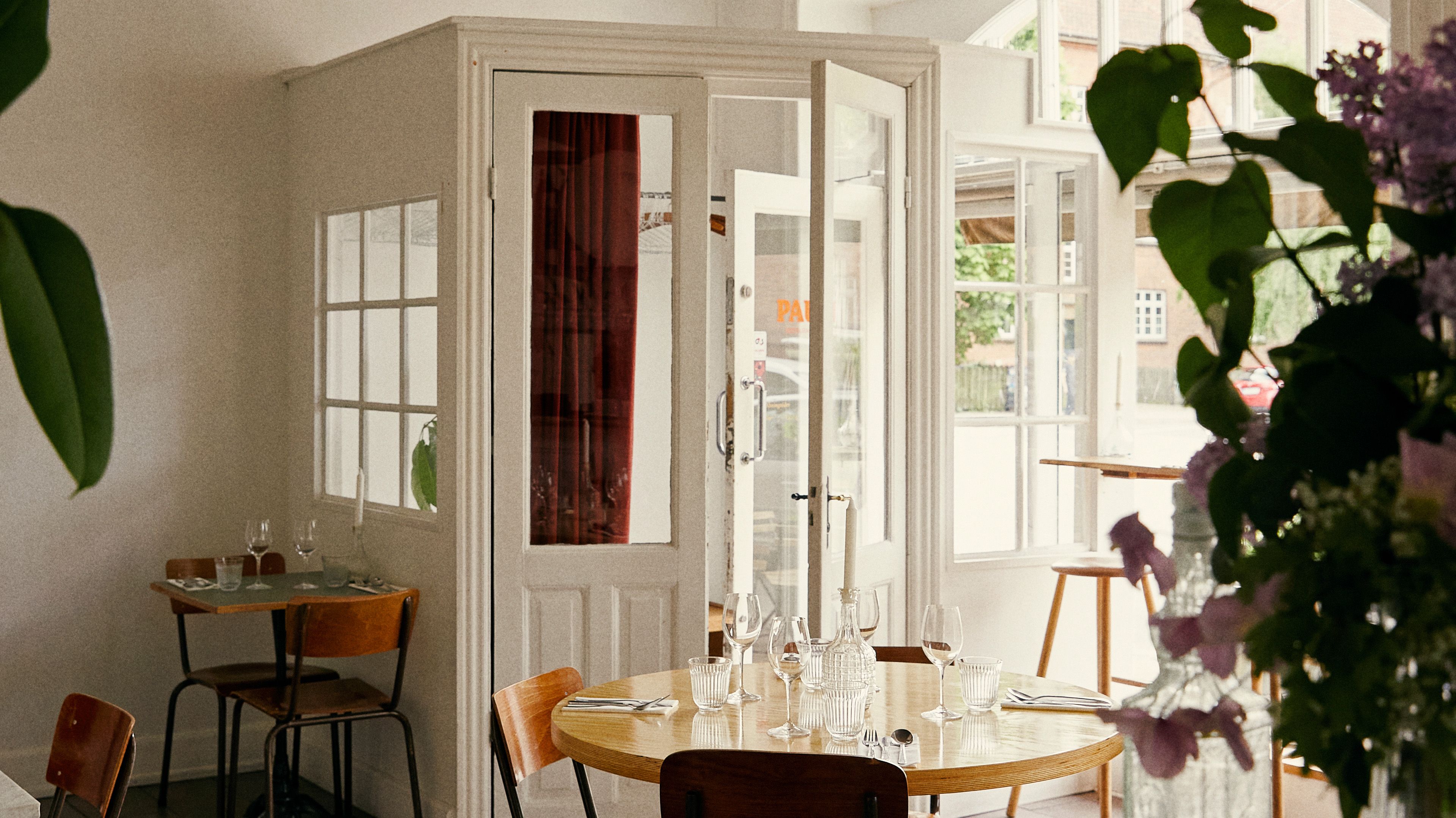 Lyst restaurant med store ruder og spisesteder indendørs. Her er flere borde med glas og krystallen lysestager. En blomster i vase står på bordet og en klippesten er på gulvet