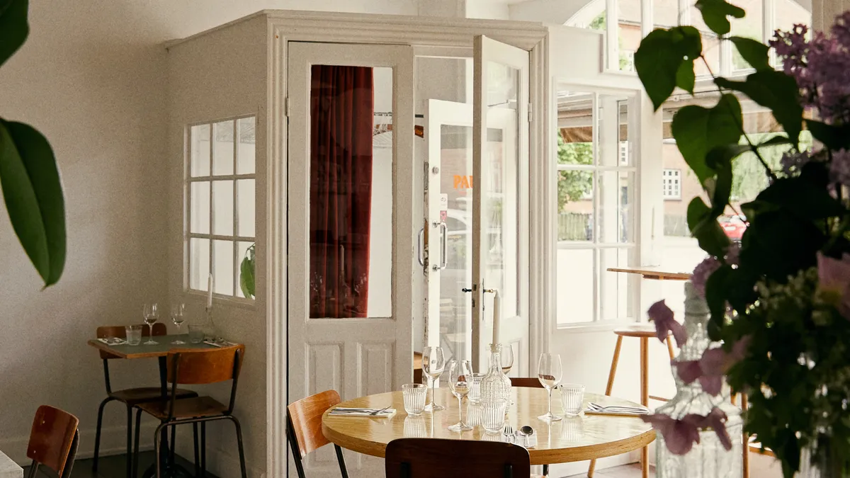 Lyst restaurant med store ruder og spisesteder indendørs. Her er flere borde med glas og krystallen lysestager. En blomster i vase står på bordet og en klippesten er på gulvet