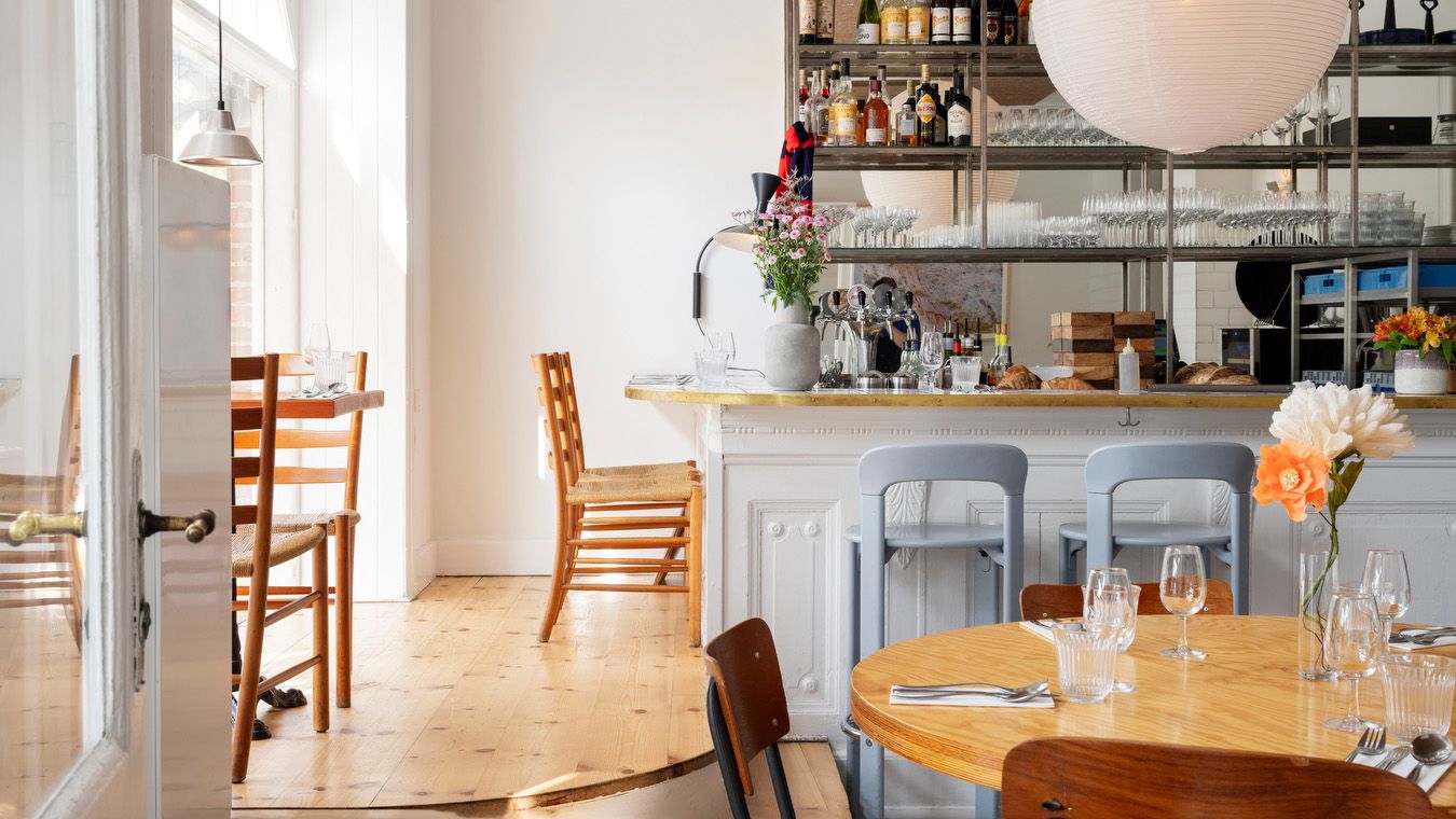 Restaurant med bord, stole, vase med blomster, vinglas og krydsfinerhyld med flasker og glas. Væg med et stort, hvidt lampe.