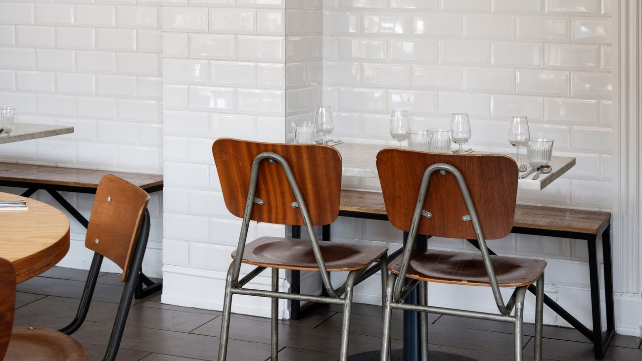 To stole med læderpuder og metalbeslag står ved et bord med glas og bestik. Væggen bag er klædt med hvide fliser og en bænk står til siden.