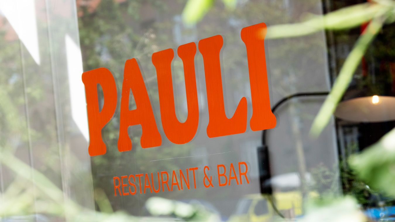 Transparente vindue med ordet Paull i orange og teksten Restaurant & Bar. Udenfor er der en grøn plante og et gult køretøj.