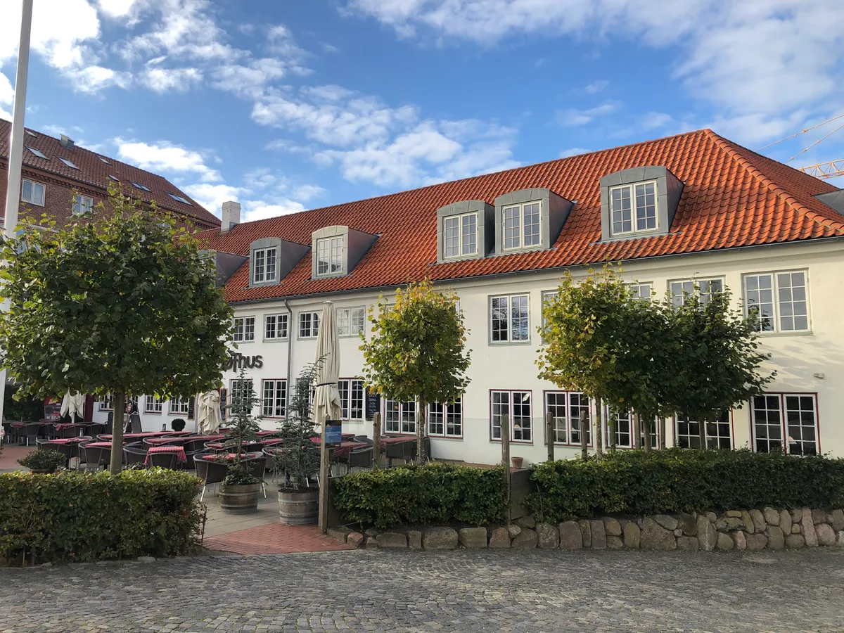 Restaurant med terrasse og udendørs møbler og parasoller. Gule røde tag med store vinduer. Træbrolægning og grønne planter. Bymidte med flere huse i baggrunden.