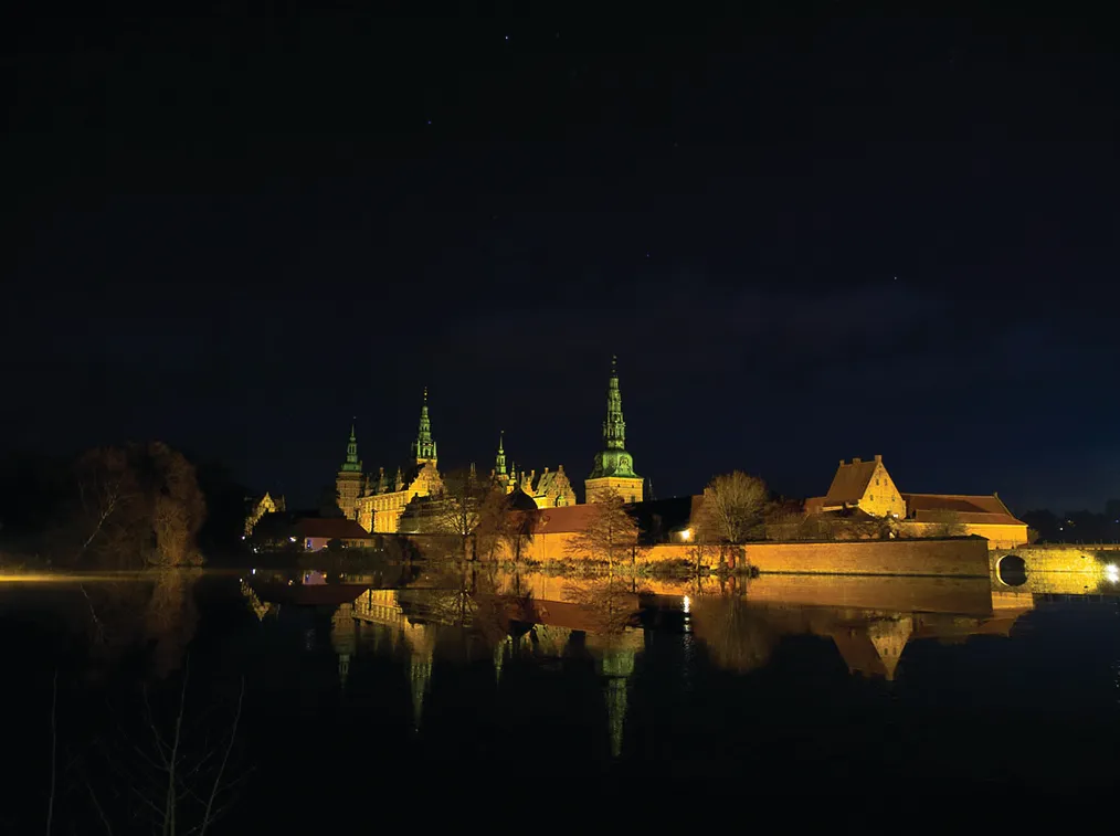 Fredagsaften ved Frederiksborg Slot med lysereflekser og tårne, der vender ud mod søen og træerne i baggrunden