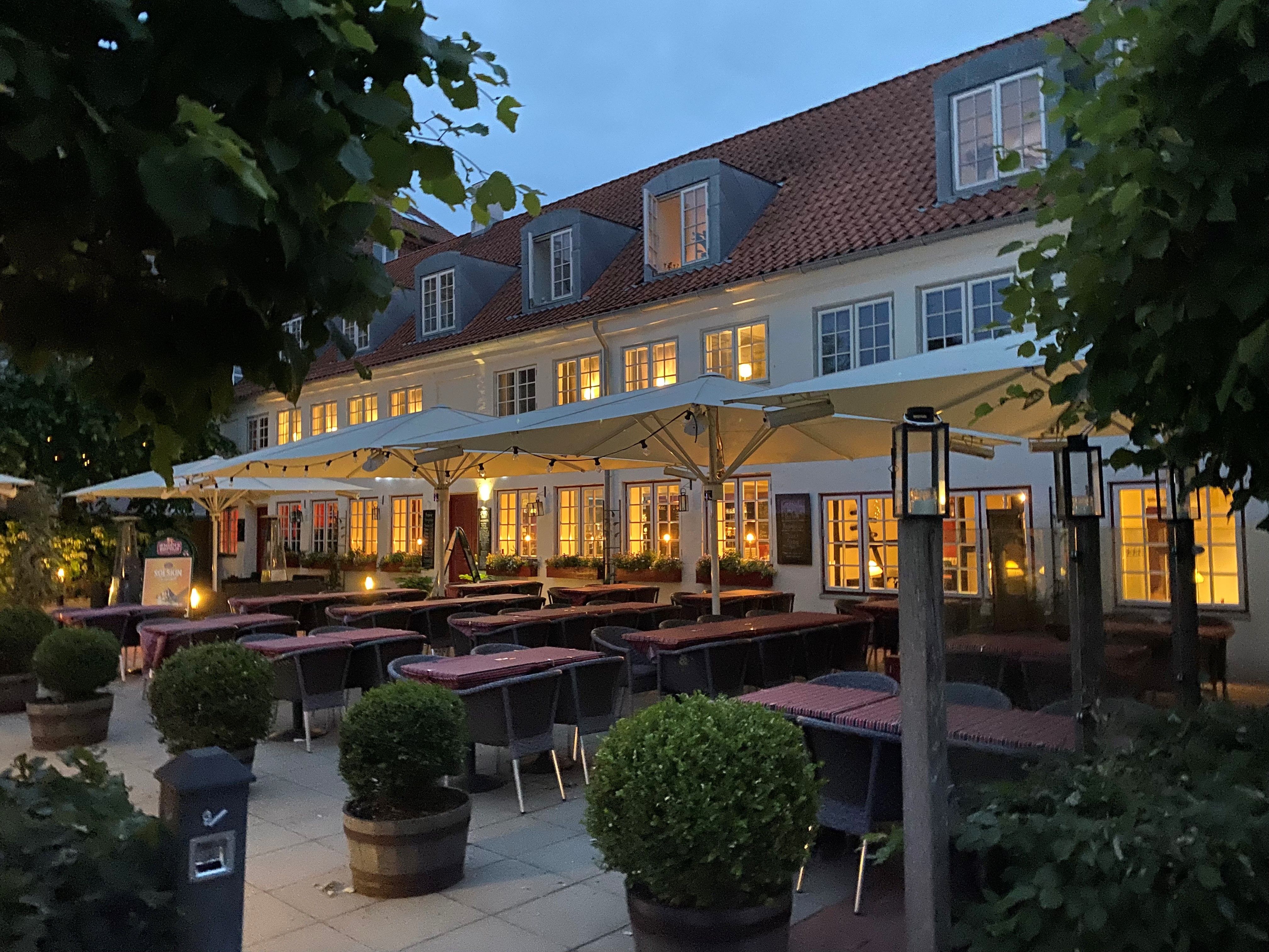 Restaurant med terrasse og træstole under parasoller, omgiven af plantepotter og lysekanoper, hvor der er mange borde med tøj, og bagved er et hus med røde tag og lyse vinduer