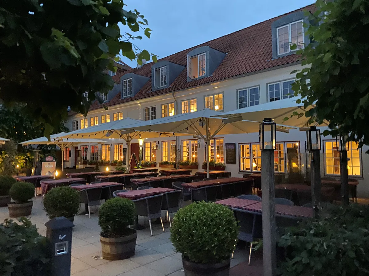 Restaurant med terrasse og træstole under parasoller, omgiven af plantepotter og lysekanoper, hvor der er mange borde med tøj, og bagved er et hus med røde tag og lyse vinduer