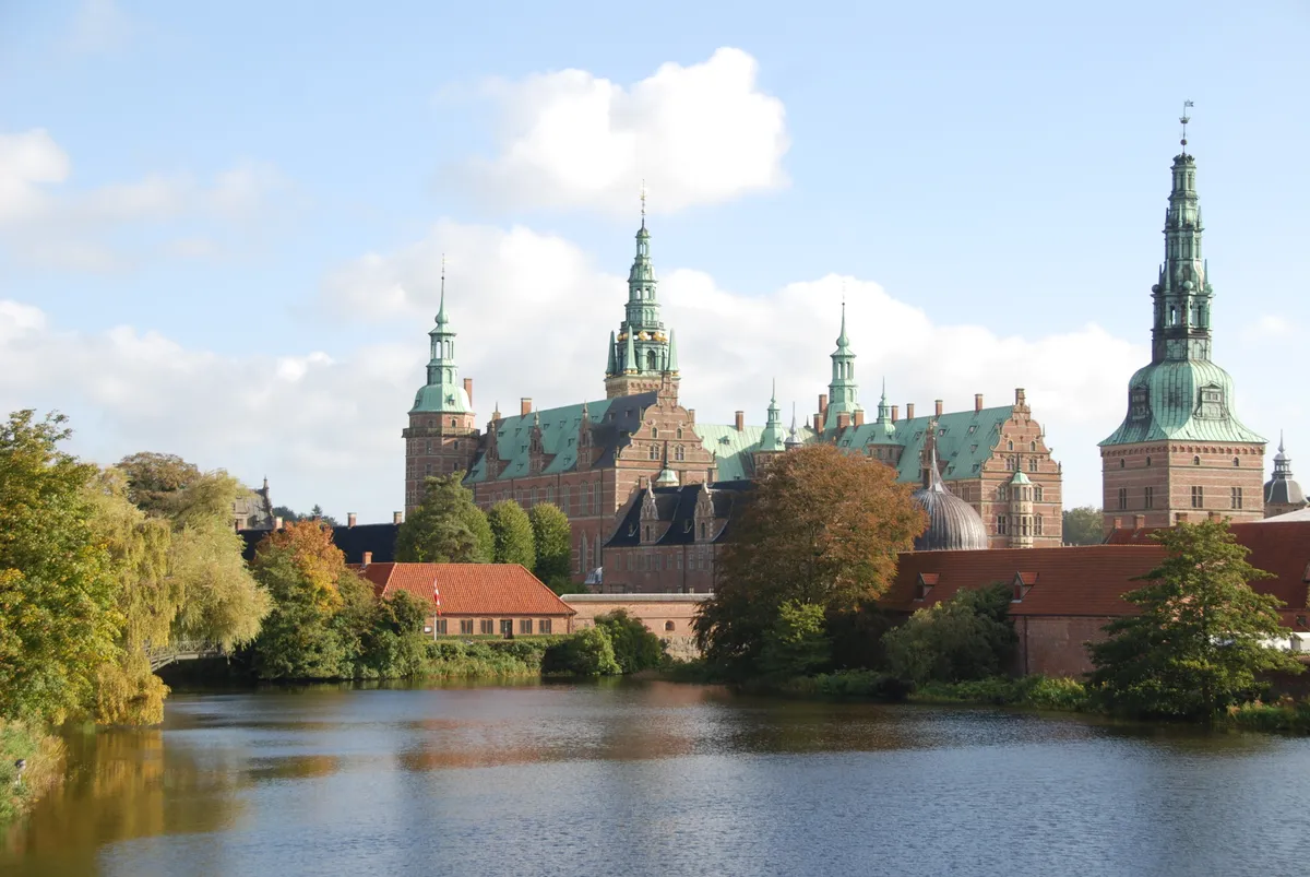 Frederiksborg Slot med grønne tag og spir, står ved en bred kanal med en smuk udsigt over slottet og lyserøde tag på bygninger i baggrunden.