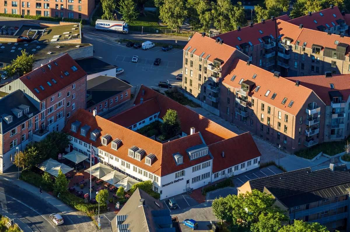 Bygninger med røde tag og mange parkeringspladser i nærheden af Slotsgade i Hillerød. Jensens Bæsthus er en restaurant med terrasse og indgang foran.
