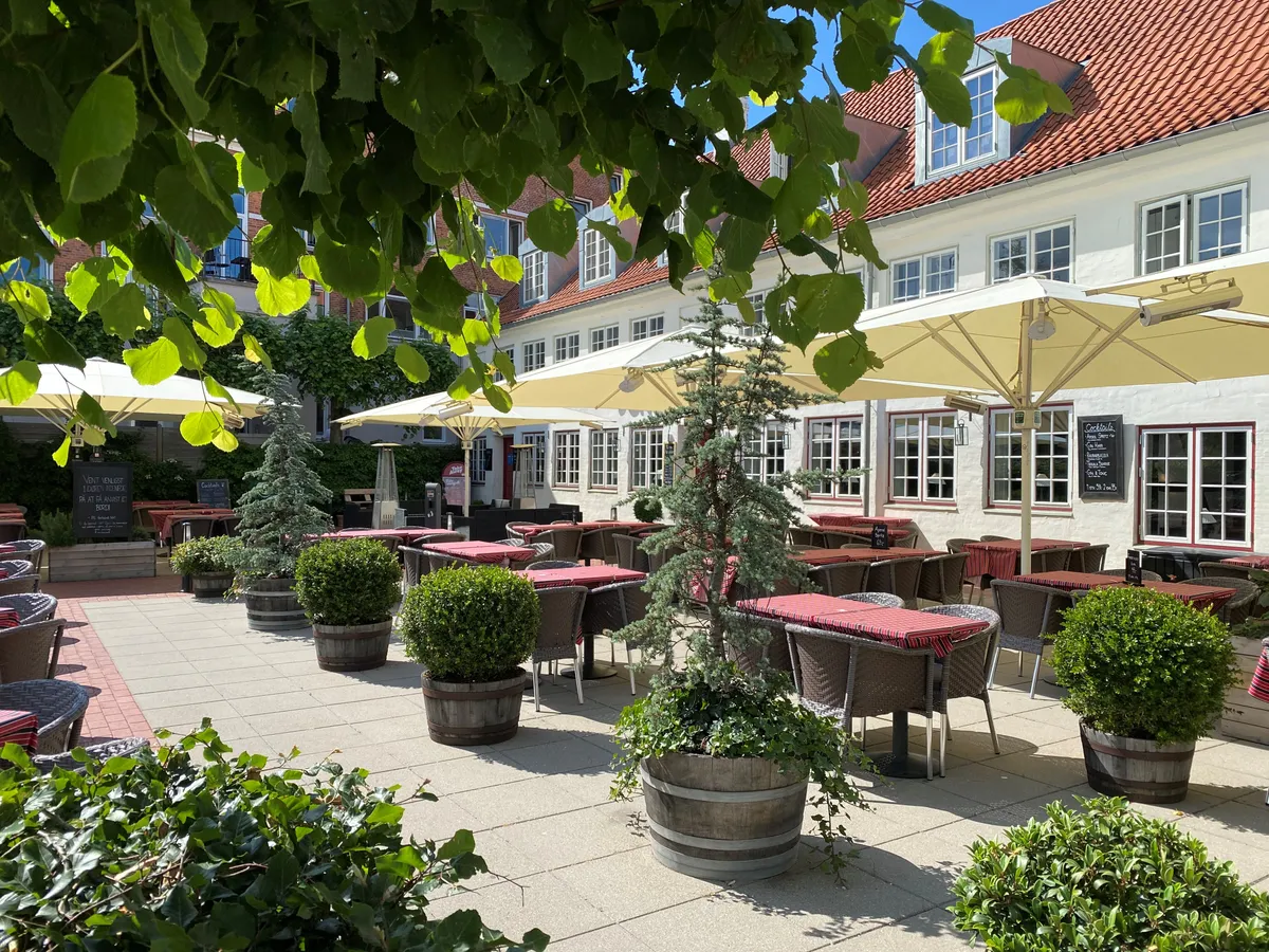 Restaurant med terrasse, møbler og parasoller i gårdhave, omgivet af grønne planter og buske, med lyserøde tag på bygningen og flere vinduer