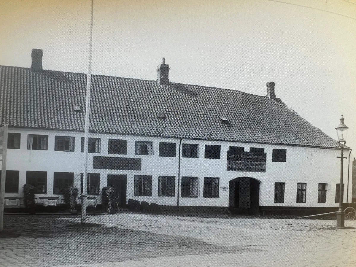 Store, lyse vinduer, to røgkammer og indgangsportal med skilt, krydsende gågade, byggede og udstyrsobjekter og søjle med gadelamper