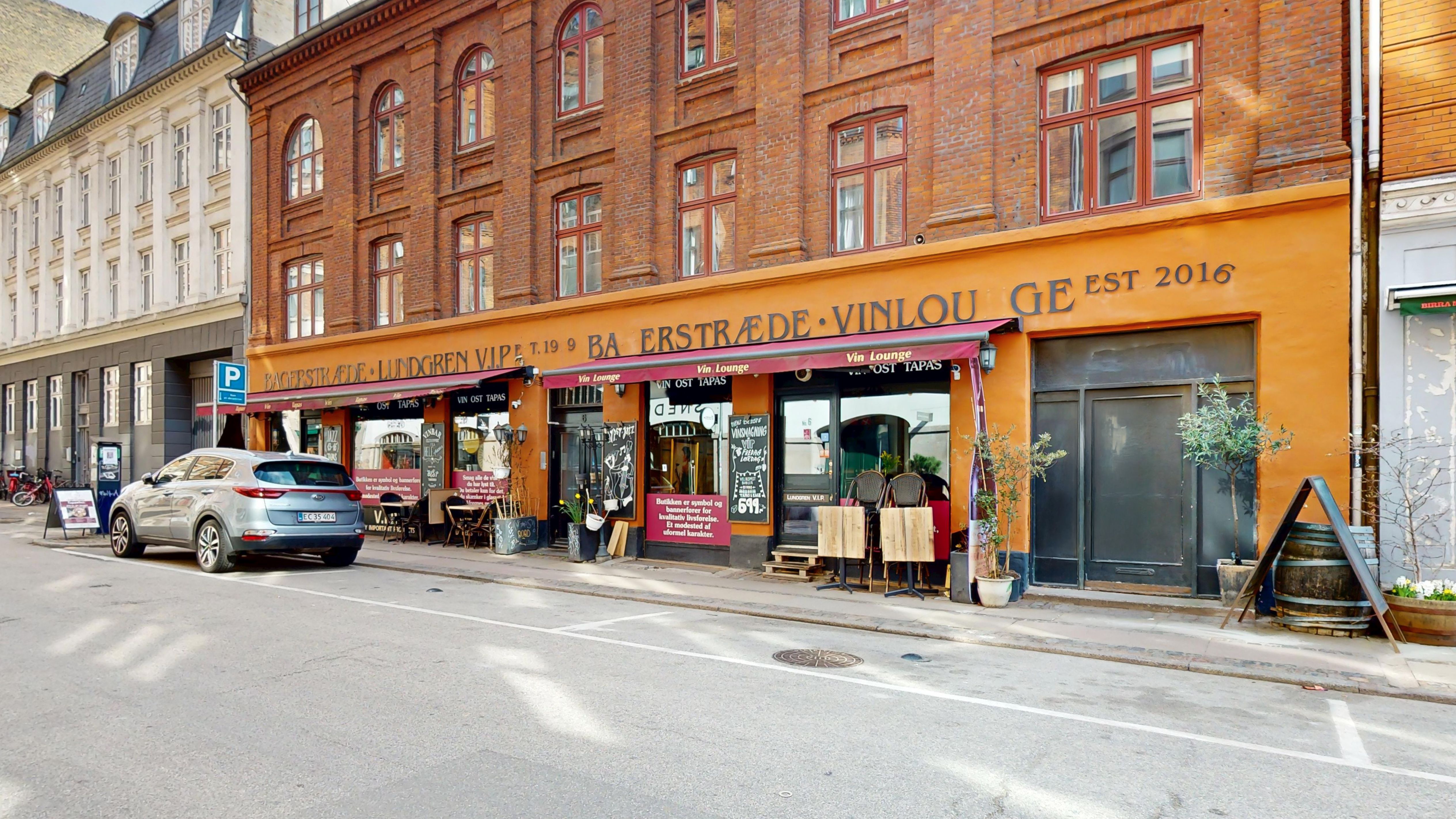 En bil er parkeret i gaden foran en vinbar med røde mærkater, og der er en række grønne planter og træer udenfor.