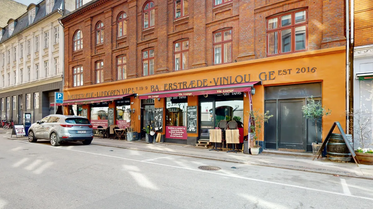En bil er parkeret i gaden foran en vinbar med røde mærkater, og der er en række grønne planter og træer udenfor.