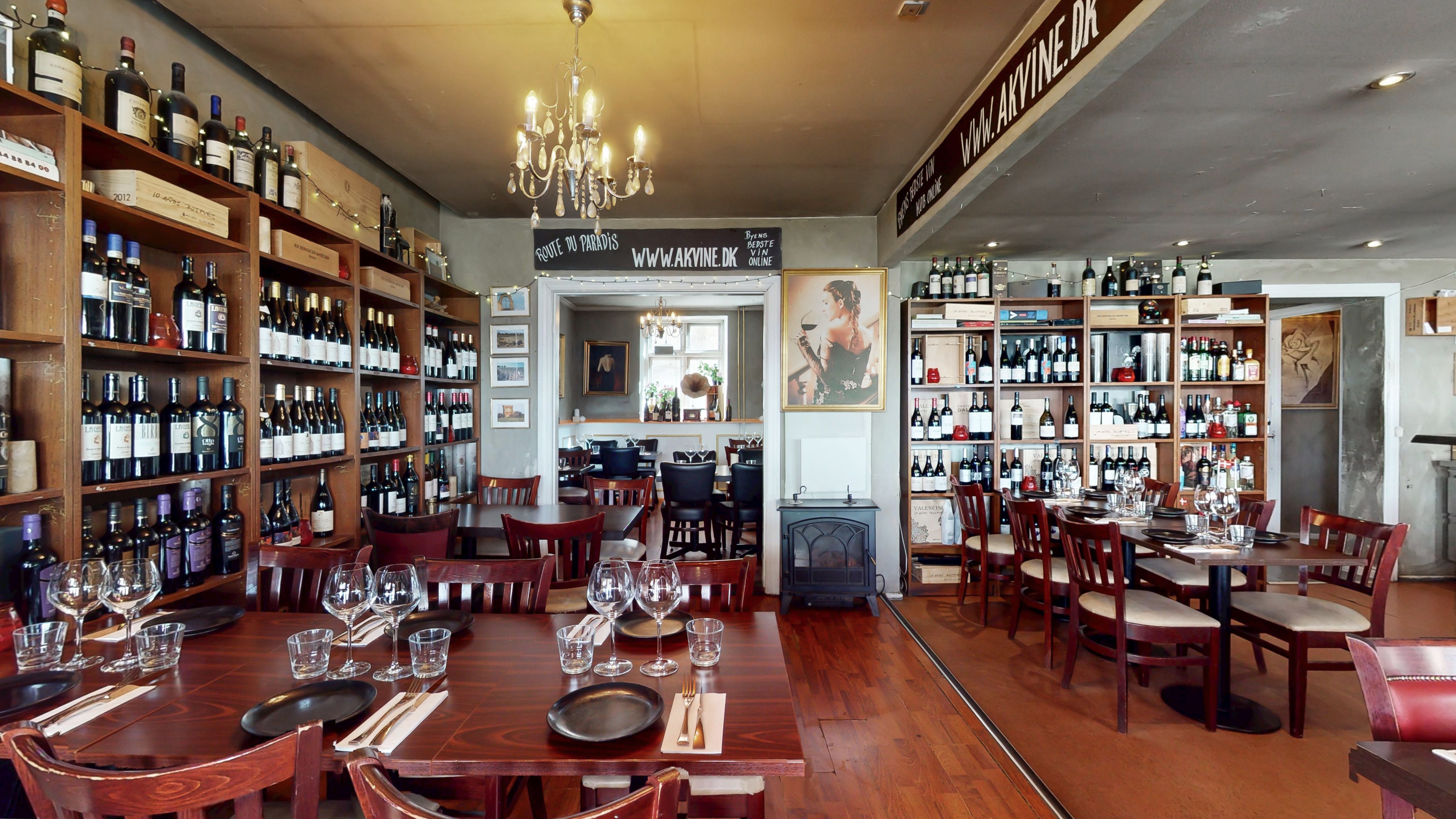 Vinbutik med mange flasker og glas samt store hylder. Væggen er beskrevet med "Route du Paradis" og "www.akvine.dk". Der er borde og stole samt lysekroner.