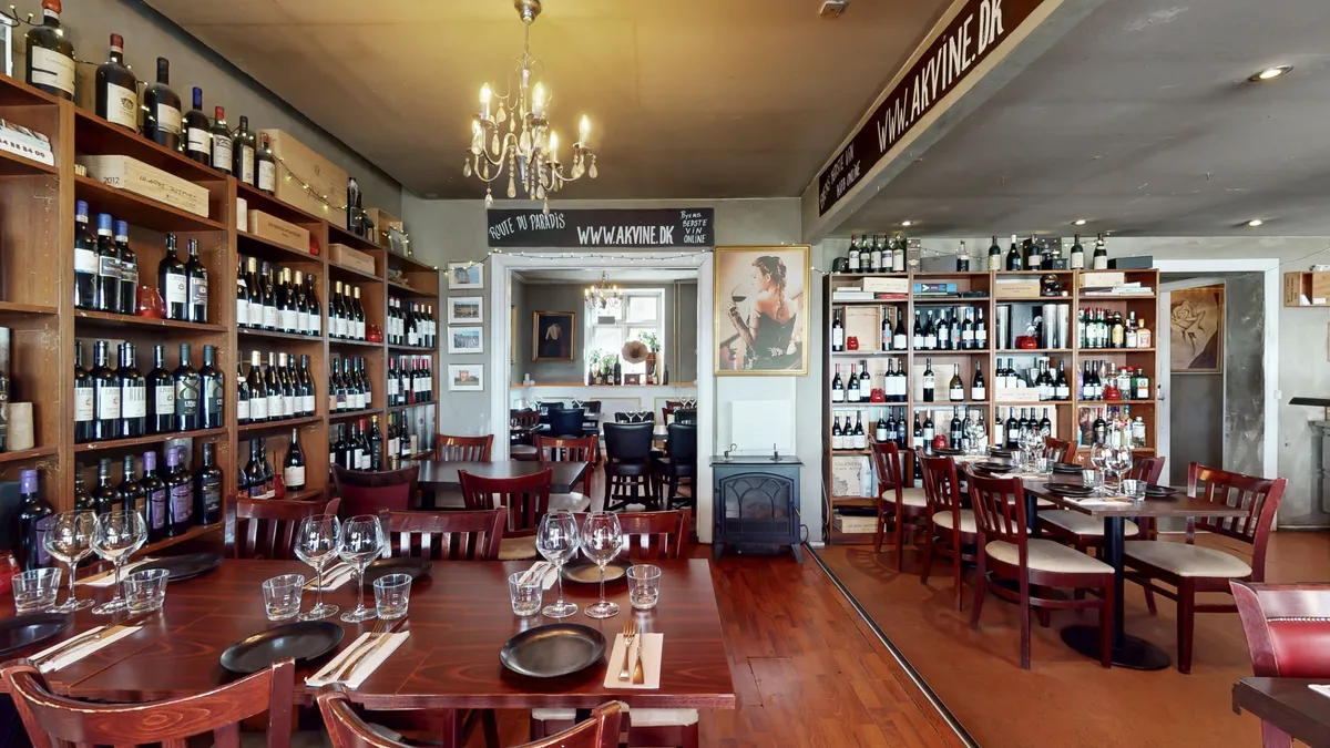 Vinbutik med mange flasker og glas samt store hylder. Væggen er beskrevet med "Route du Paradis" og "www.akvine.dk". Der er borde og stole samt lysekroner.
