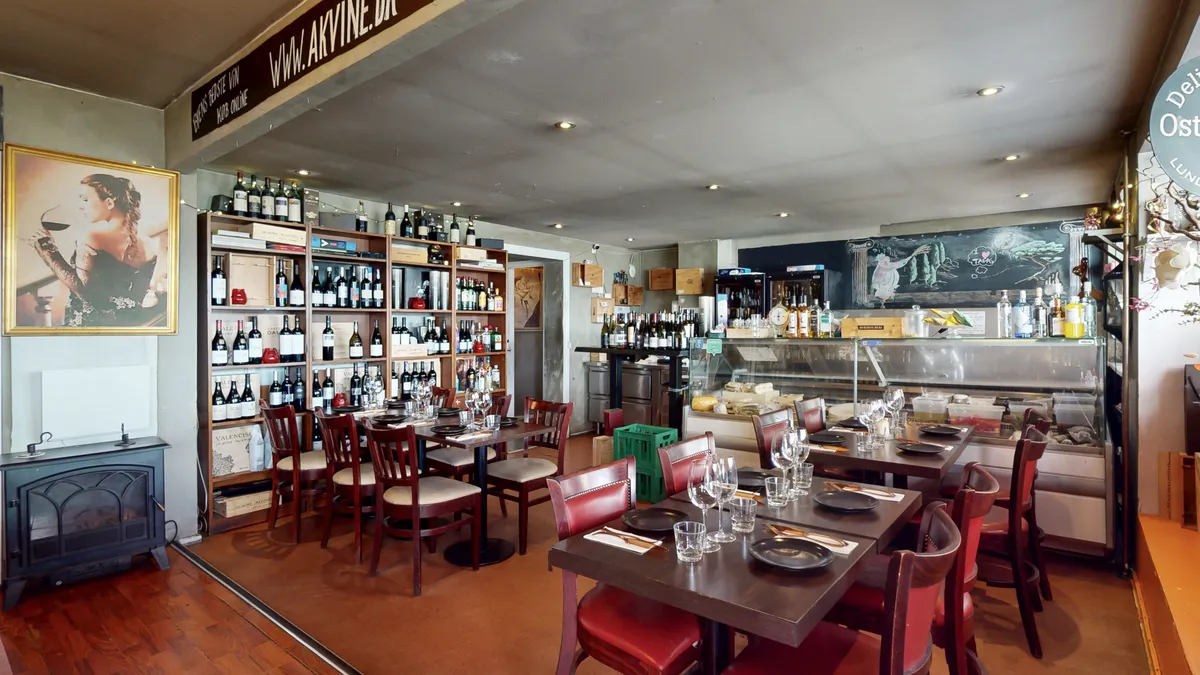 Køkken med borde og stole, vinflasker og øl på hylder og sælger drikkevarer bag baren.